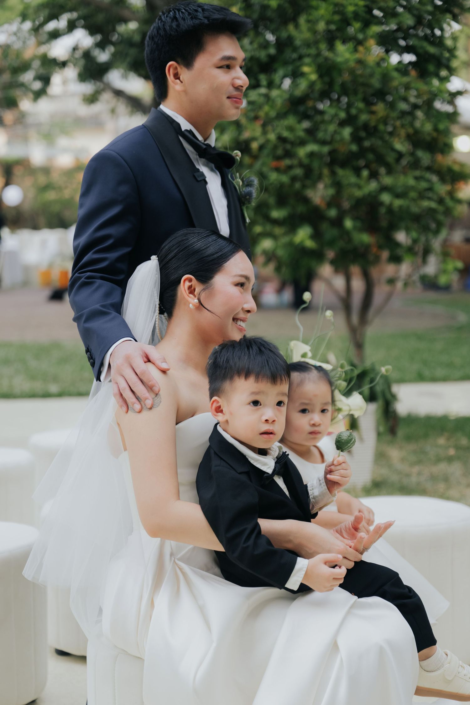 Asian bride and groom in wedding attire sit outdoors with two young children dressed in formal wear.