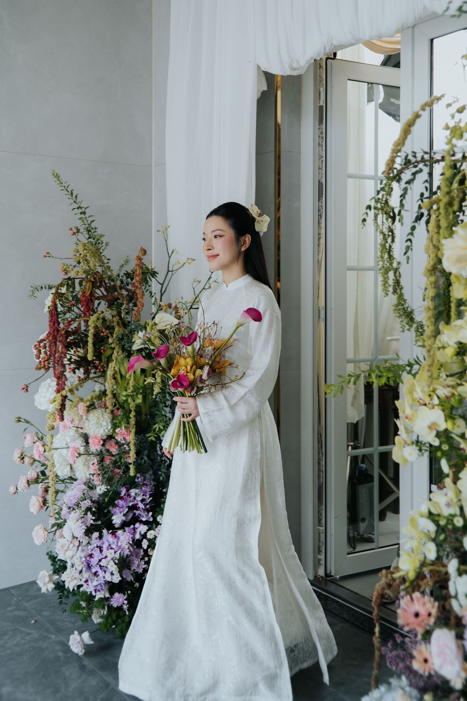 Radiant Vietnamese bride in white ao dai holding a colorful wildflower bouquet standing beside lush floral arrangements.