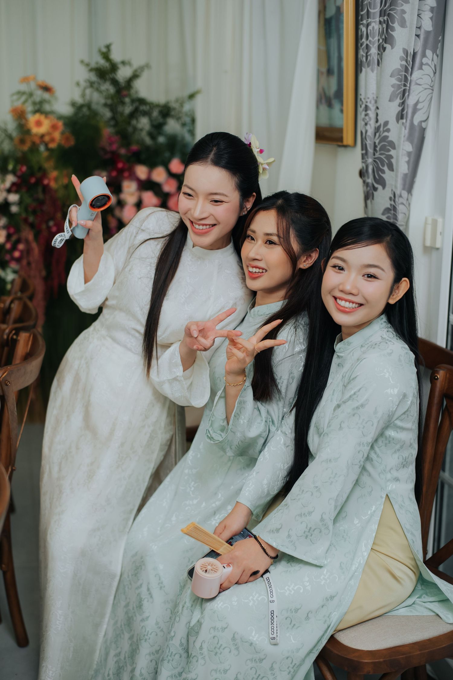 Three young women in matching white ao dai dresses smiling and posing together at a traditional Vietnamese celebration.