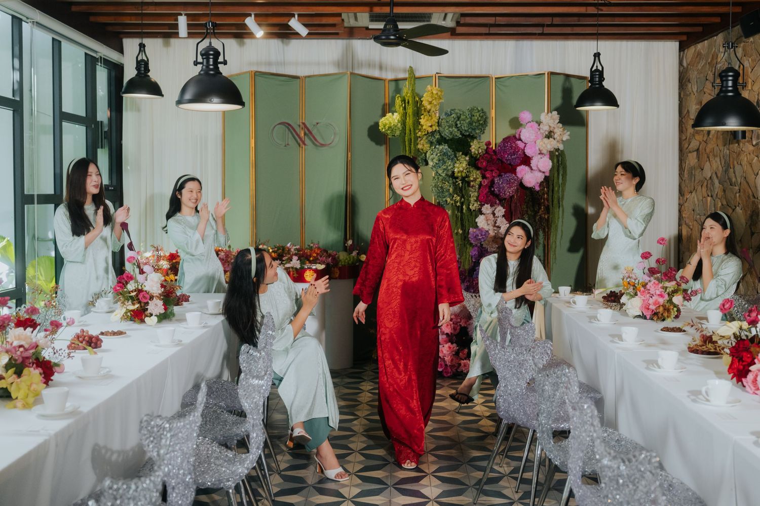 Man in red ao dai walks between bridesmaids in gray dresses at a floral Vietnamese wedding reception.