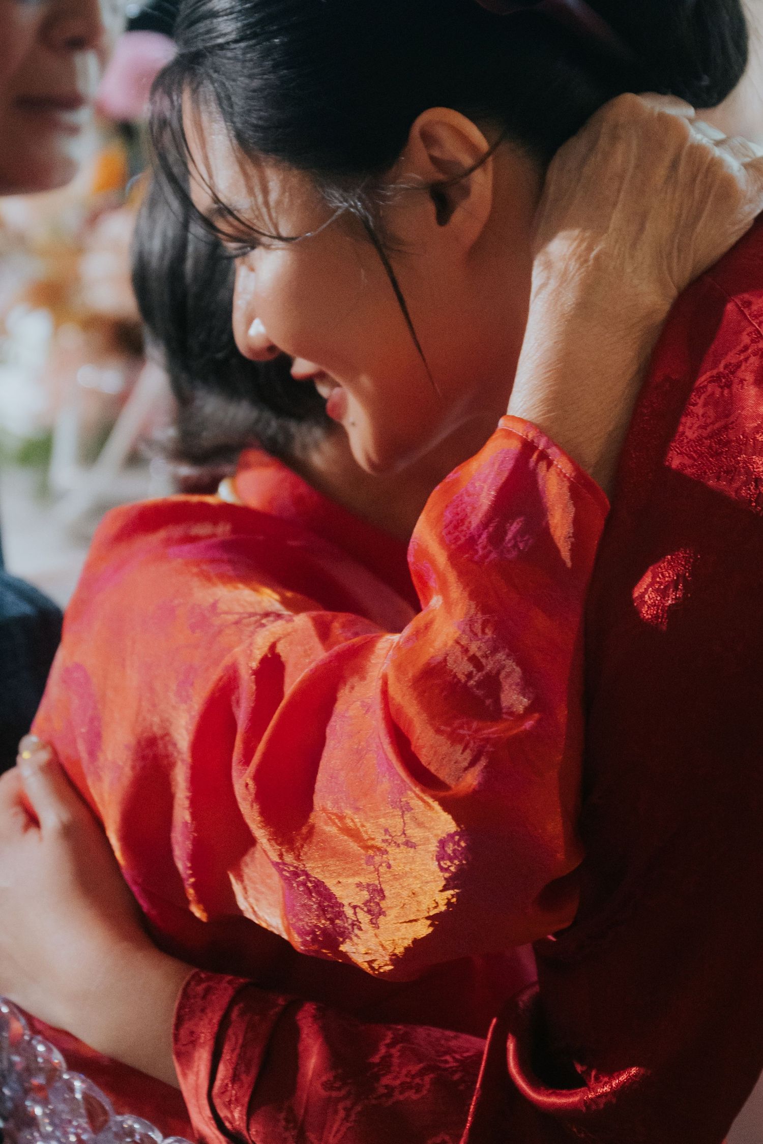 Two women in red traditional Chinese attire share a warm embrace at a celebratory event.