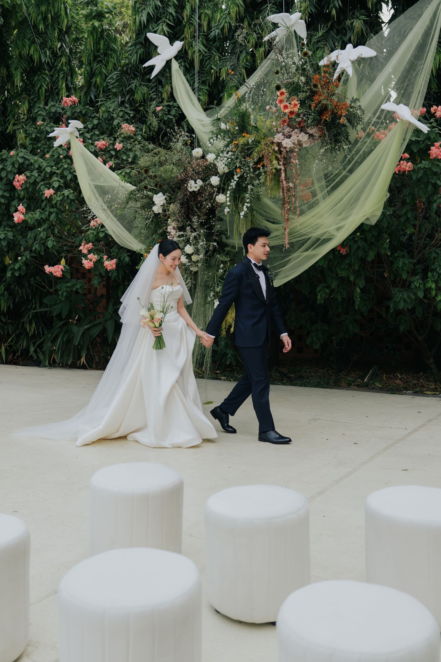 Newlyweds walk hand-in-hand past white doves and floral greenery wall at elegant outdoor wedding.