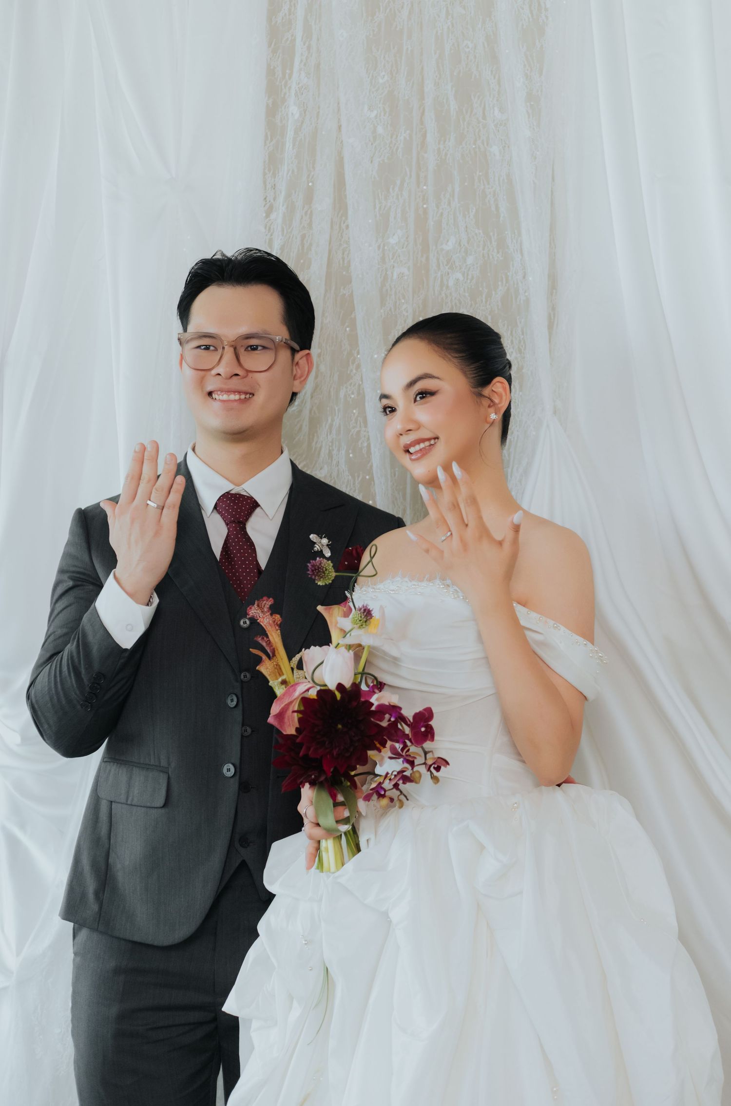 Newlywed couple in wedding attire showing rings, groom in dark suit, bride in white gown holding burgundy bouquet.