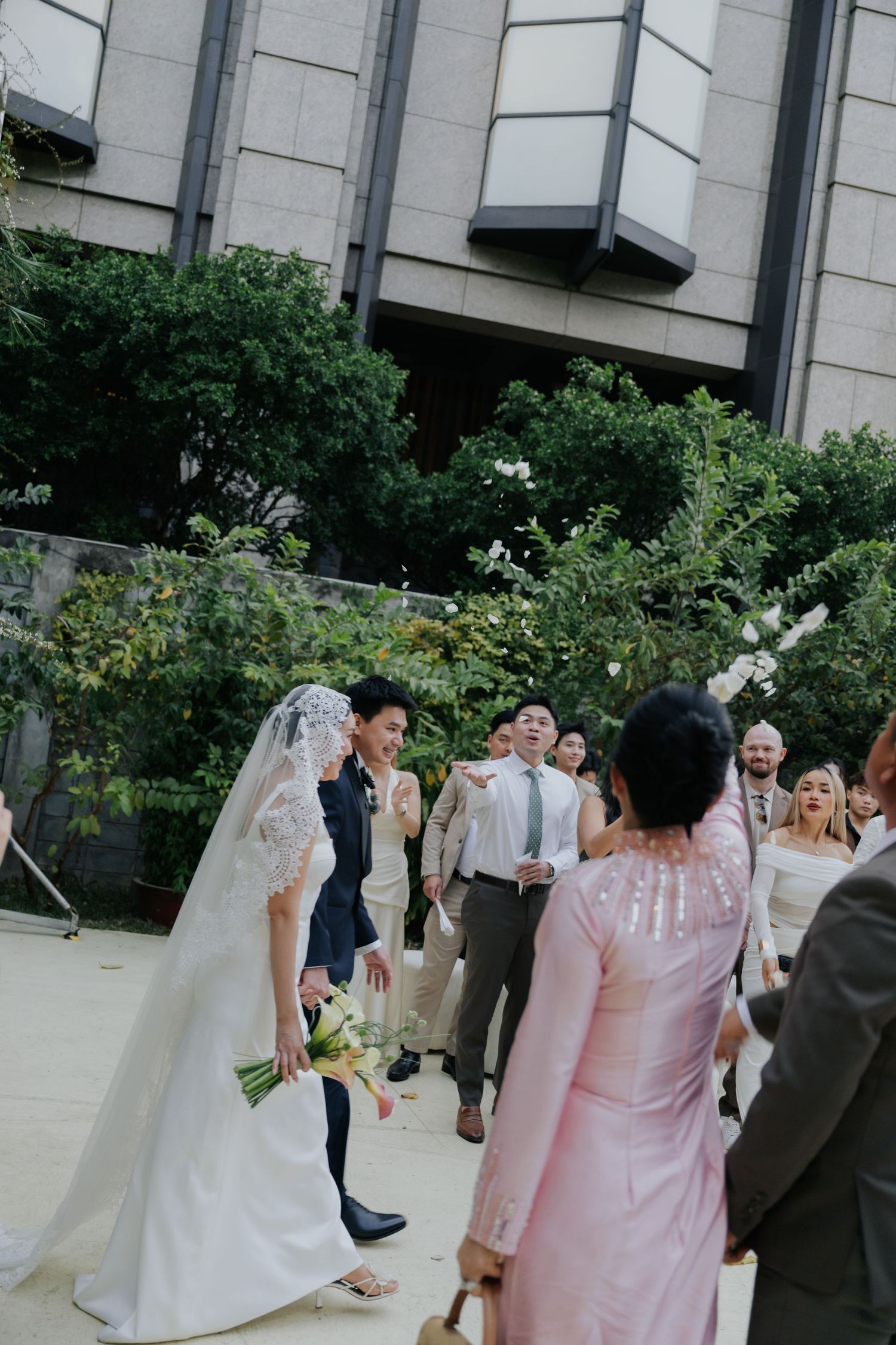 Bride in white gown and veil holds bouquet while greeting guests outdoors at an elegant wedding venue.