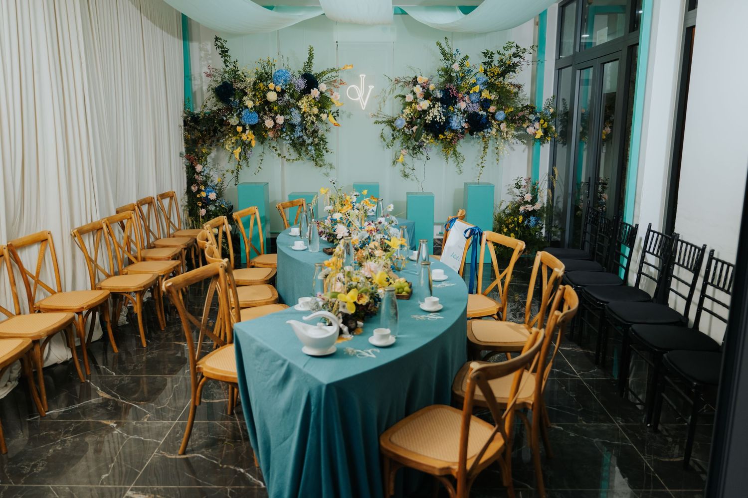 Elegant wedding reception with teal tablecloths, wooden cross-back chairs, and floral arrangements in blue and yellow tones.
