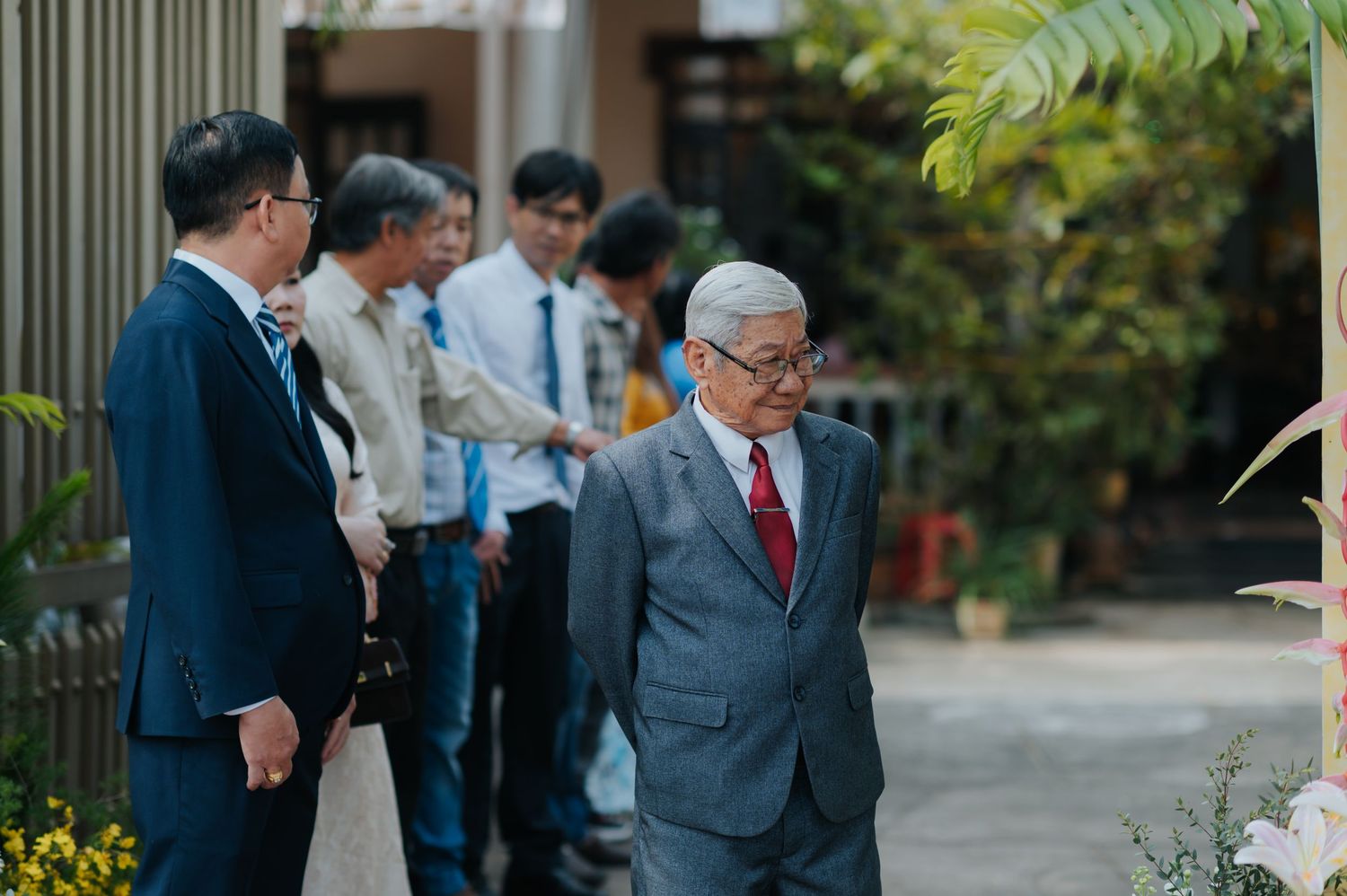Elderly man in gray suit smiling outdoors, walking with group of men in formal attire near tropical garden.