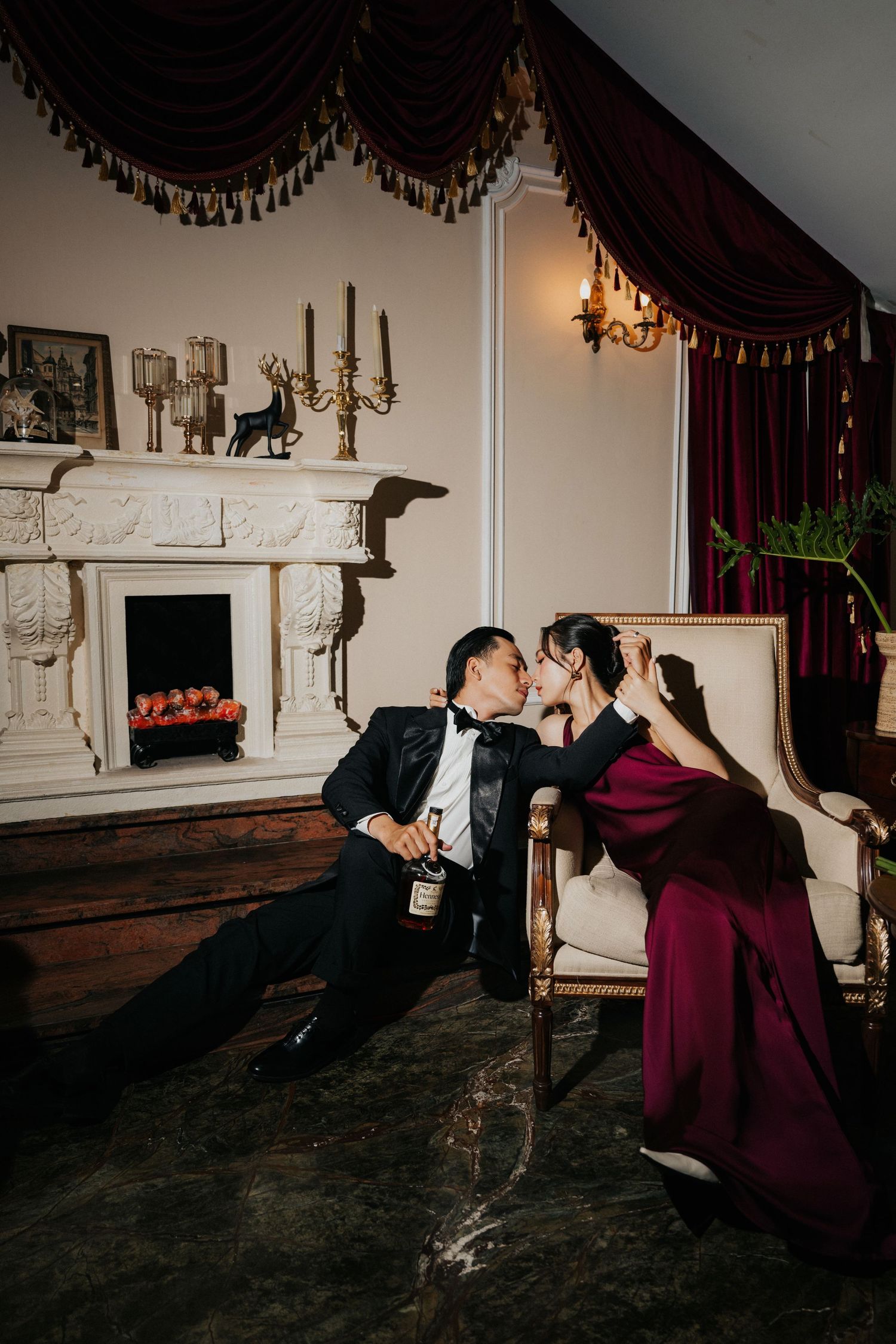 Elegant couple in formal attire sharing a romantic moment by a fireplace in a luxurious room with burgundy drapes.