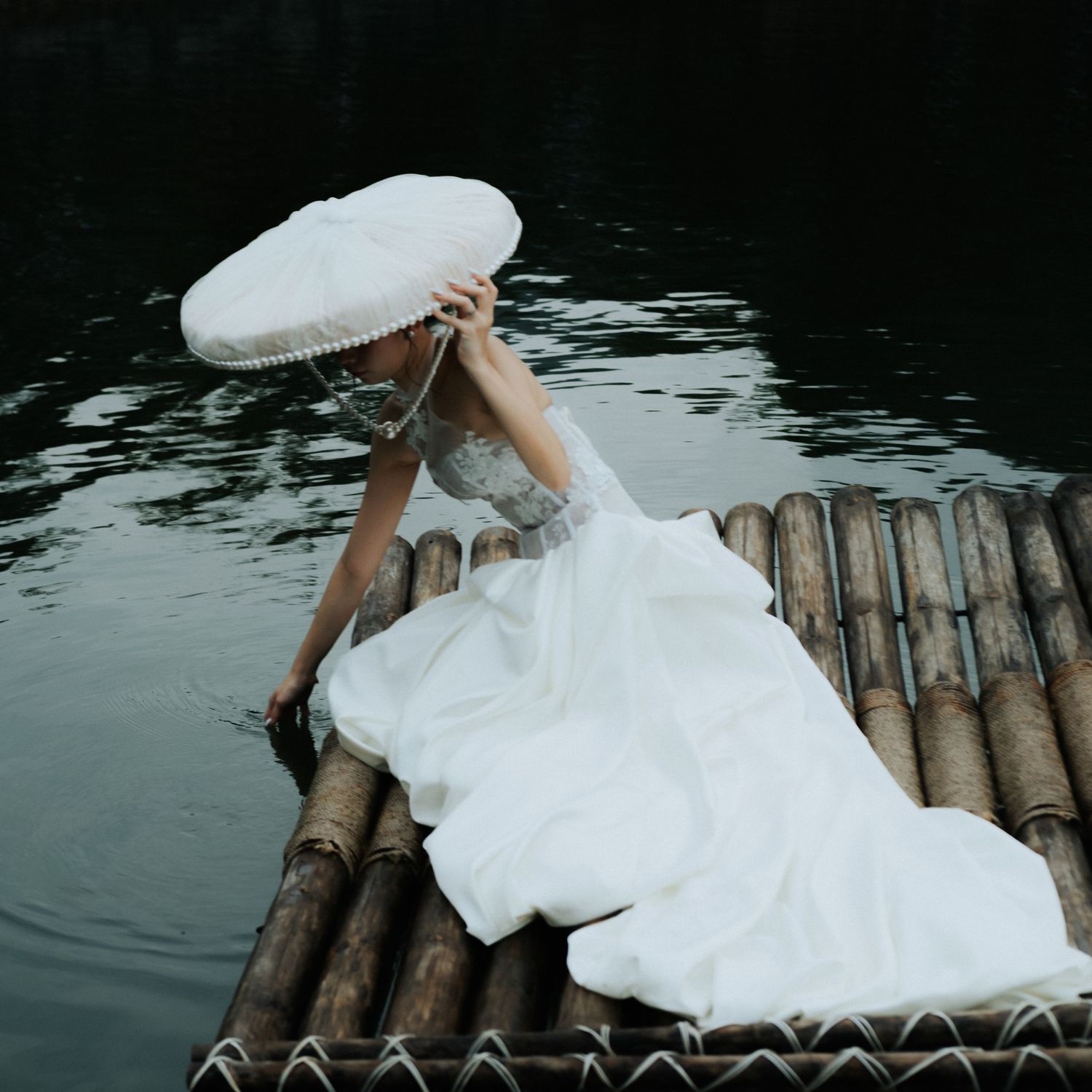 A bride in a white gown sits on a bamboo raft by dark water, holding a white parasol umbrella.