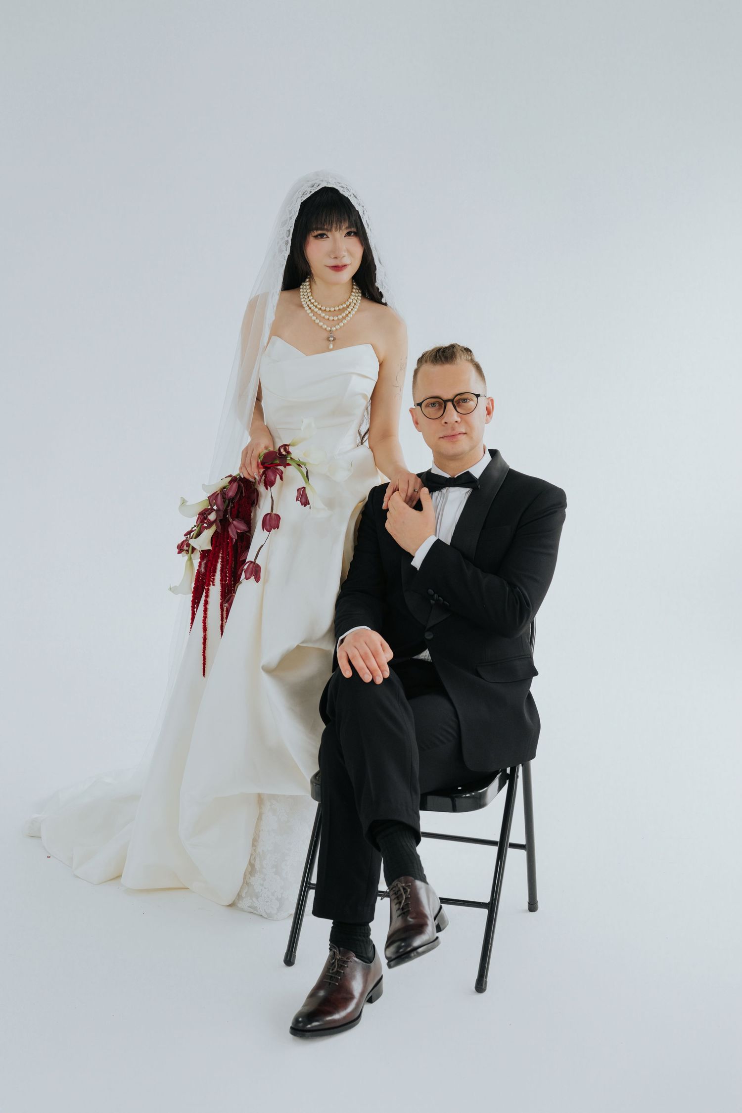 Bride in white gown holding burgundy bouquet stands beside groom in black tuxedo seated on chair against white background.