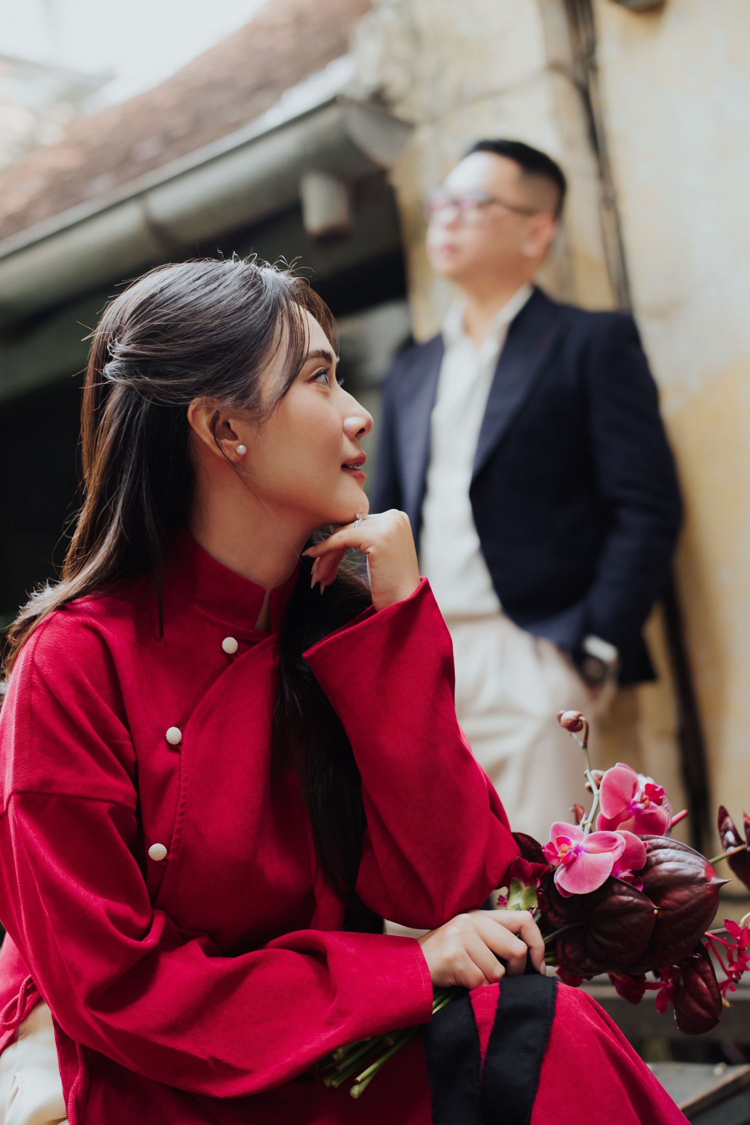 Young woman in red ao dai holding flowers with man in blue blazer standing behind her.