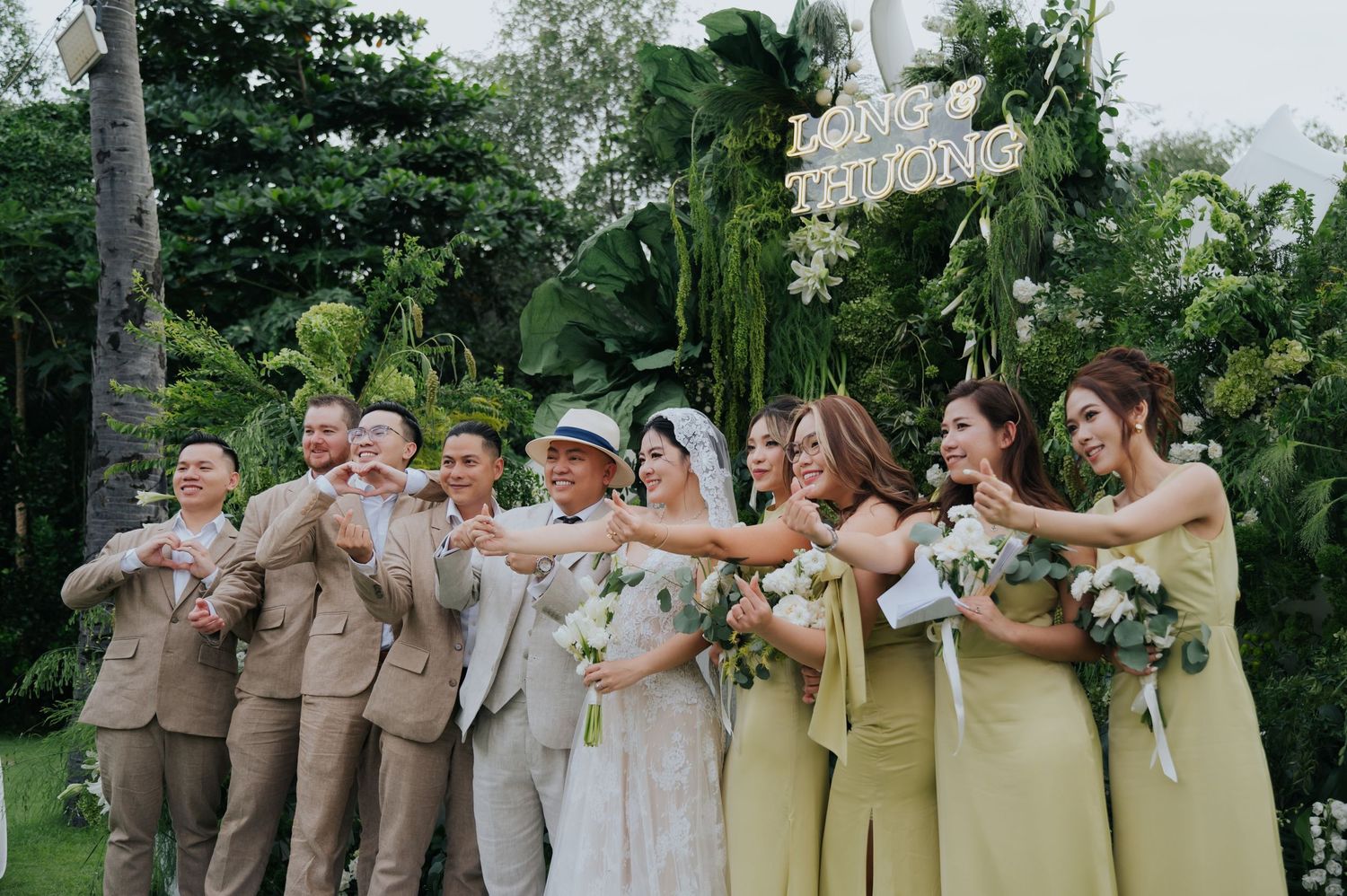 Wedding party poses outdoors with bride in white, groom, groomsmen in tan suits, and bridesmaids in yellow-green dresses.