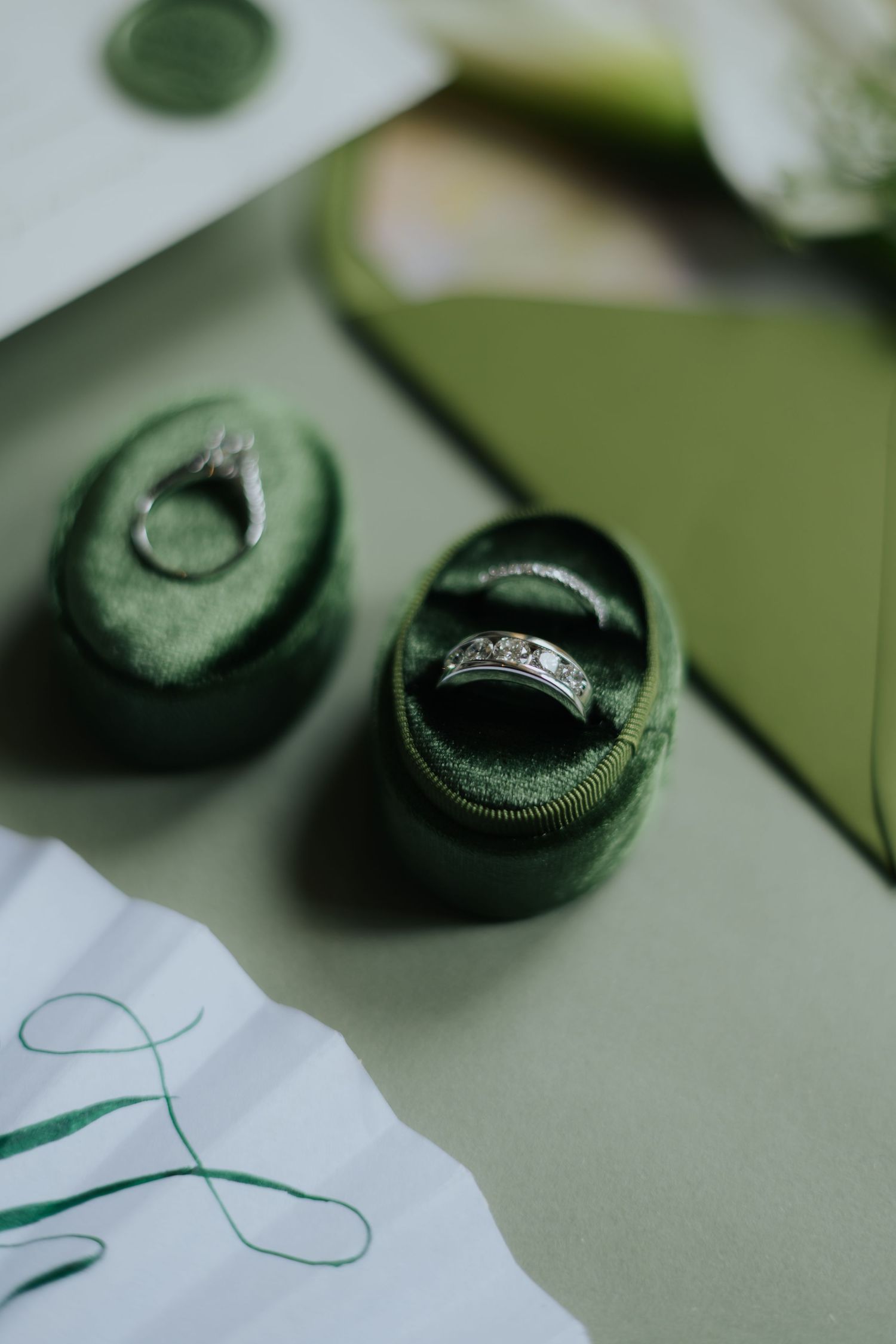 Two silver rings displayed in open green velvet ring boxes on a sage green surface with wedding stationery.