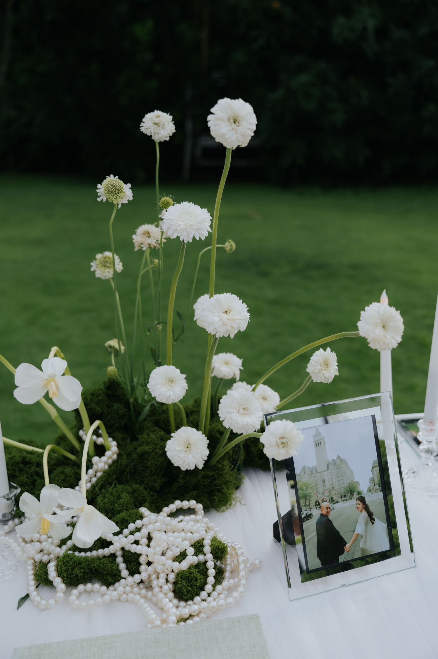 White ranunculus and moss centerpiece with pearl necklace and framed wedding photo on white tablecloth outdoors.