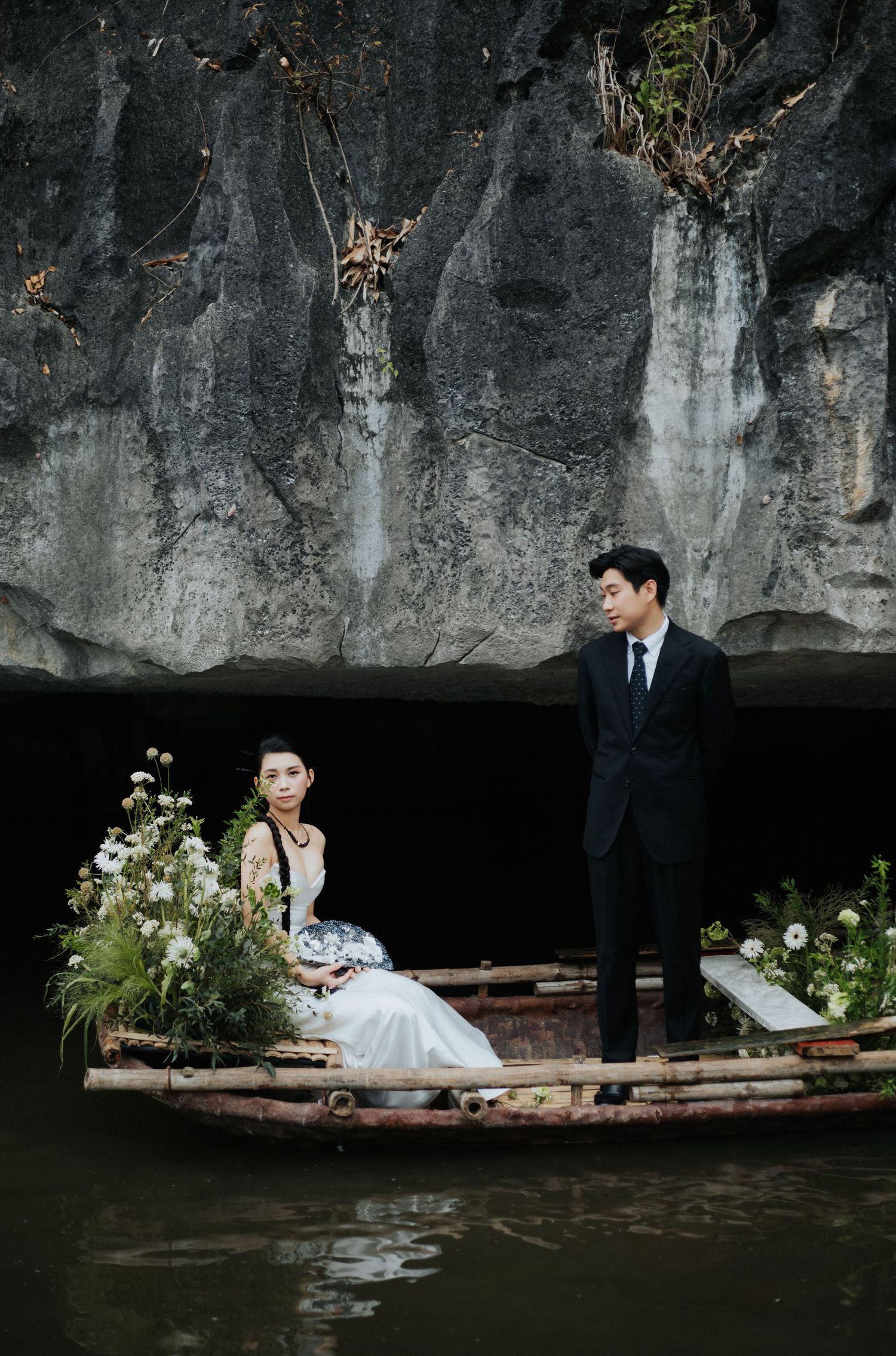 Couple in wedding attire on a flower-adorned wooden boat near a rocky cliff, bride seated among white blooms.