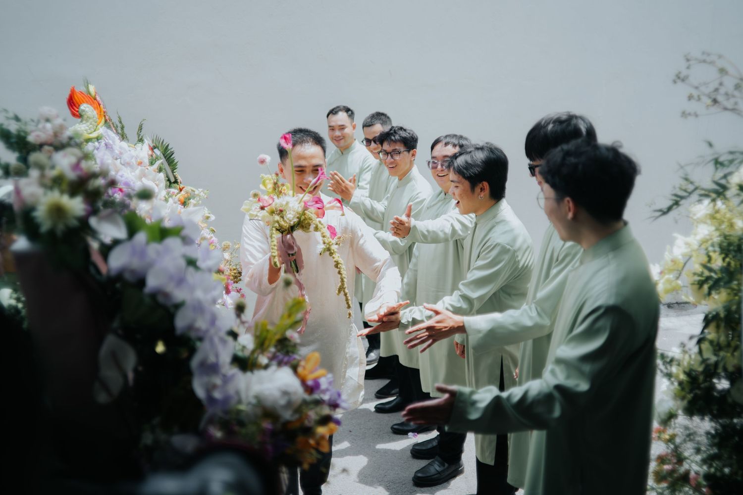 A bride in a white dress receives flowers from groomsmen in green ao dai at an elegant outdoor wedding ceremony.