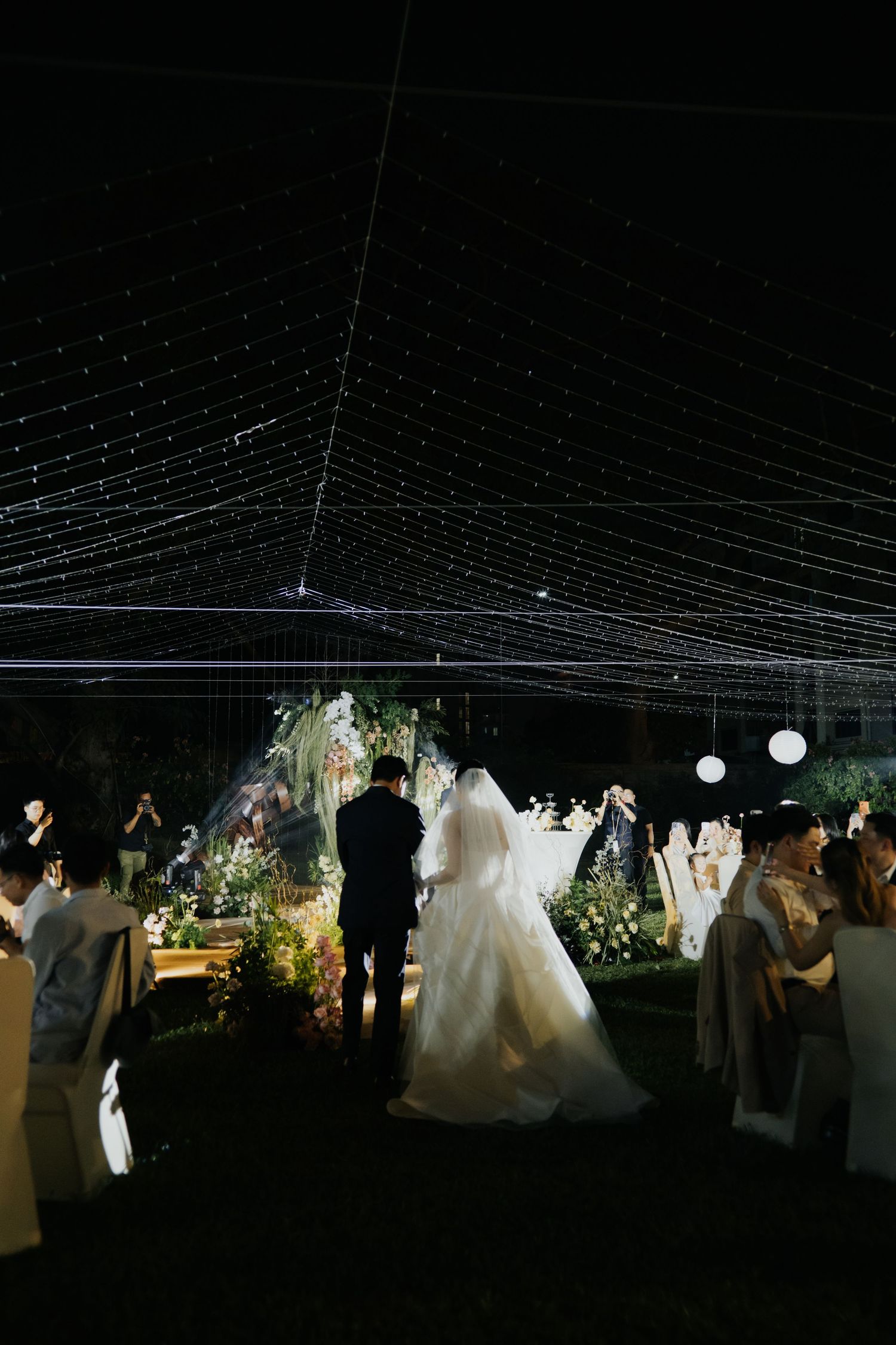 Bride and groom walk down the aisle under a canopy of twinkling string lights at an elegant outdoor night wedding.