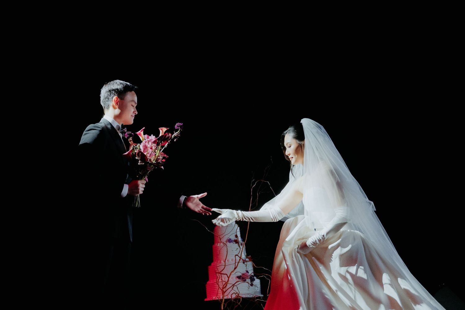 A groom presents flowers to a bride in a white wedding gown and veil against a dark background.