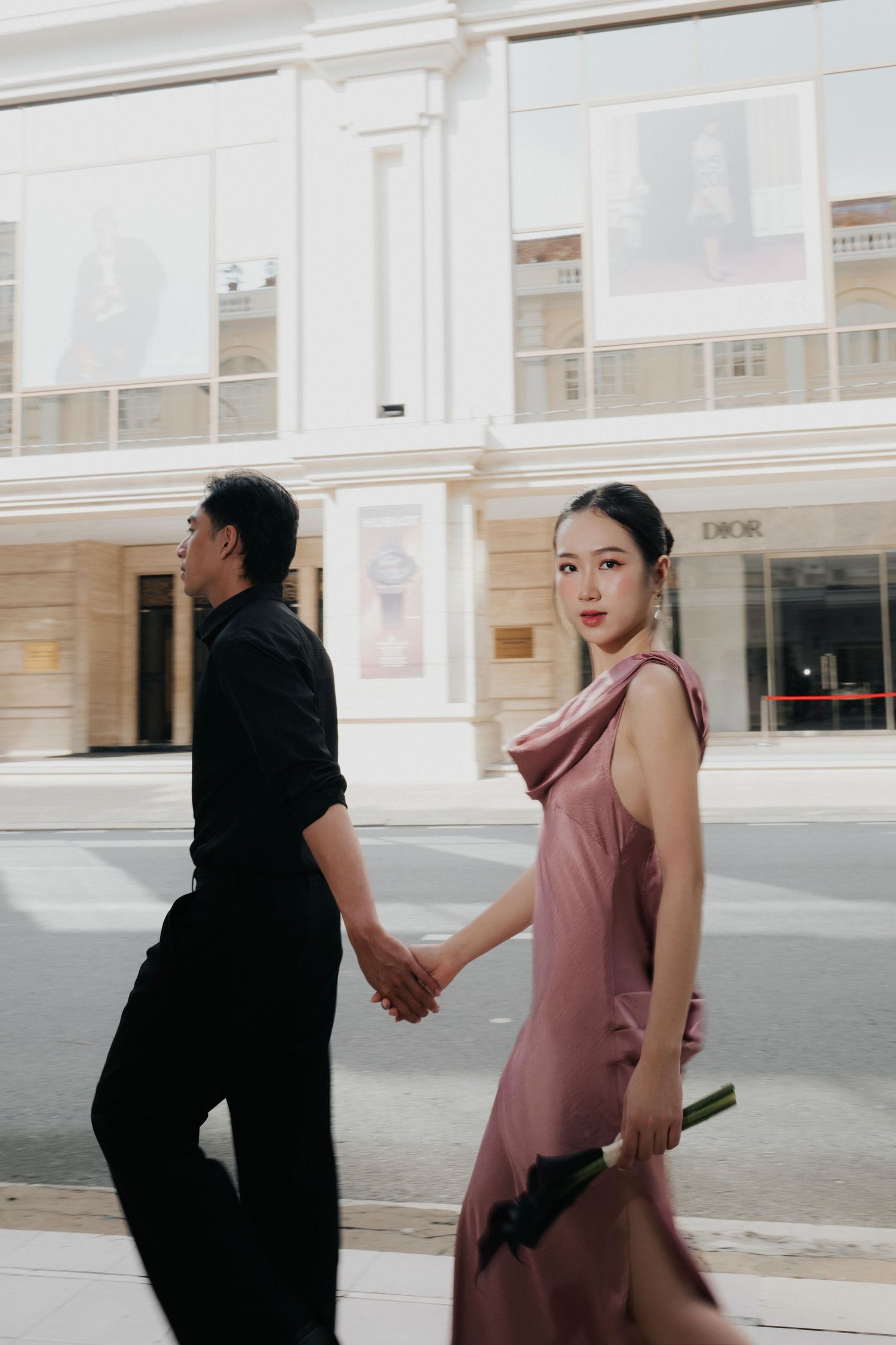 A couple holds hands walking past a Dior store, woman in elegant pink dress glancing back over her shoulder.