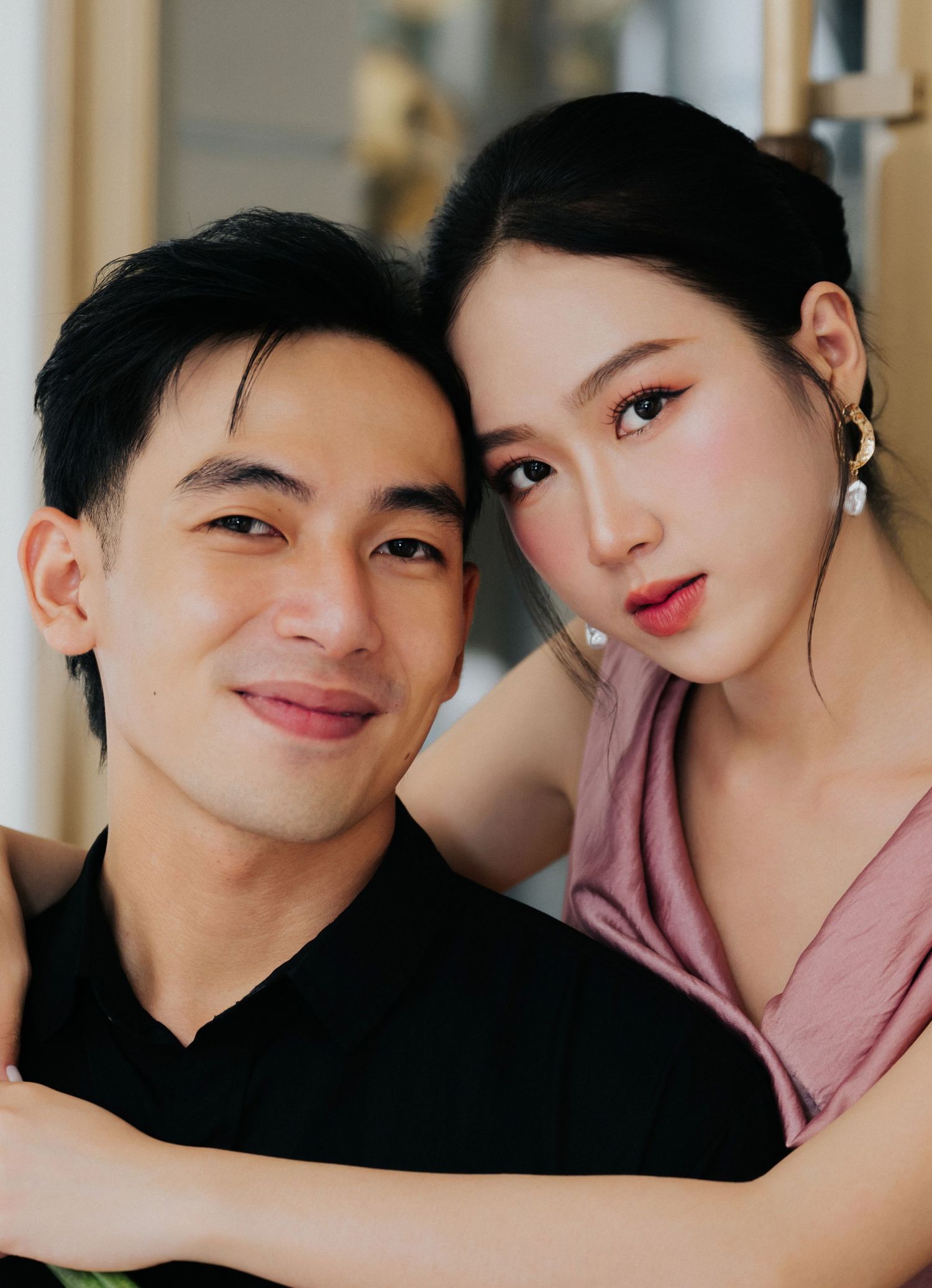 A young Asian couple poses closely together, the woman in a pink top with pearl earrings leaning on the mans shoulder.
