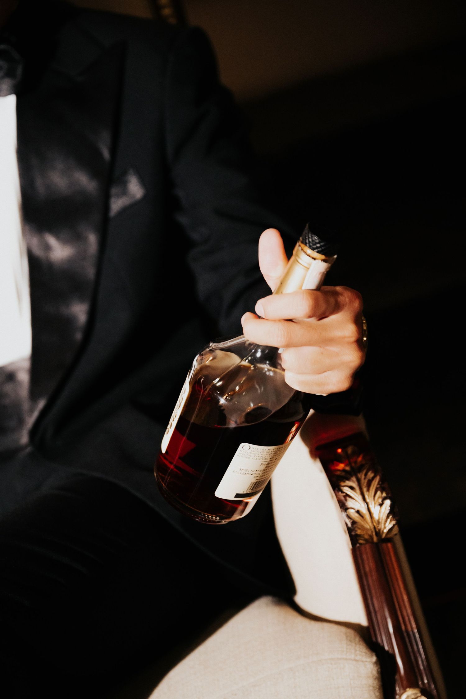 Man in black suit holding a bottle of whiskey while seated in a dimly lit room.