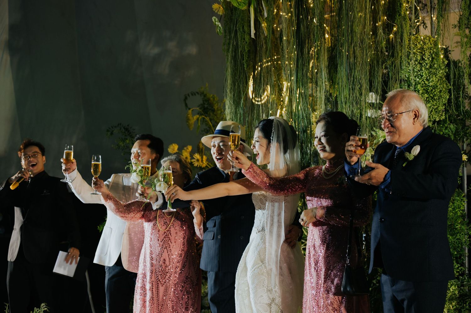 Wedding party toasting with champagne glasses at elegant reception with lush green floral backdrop.