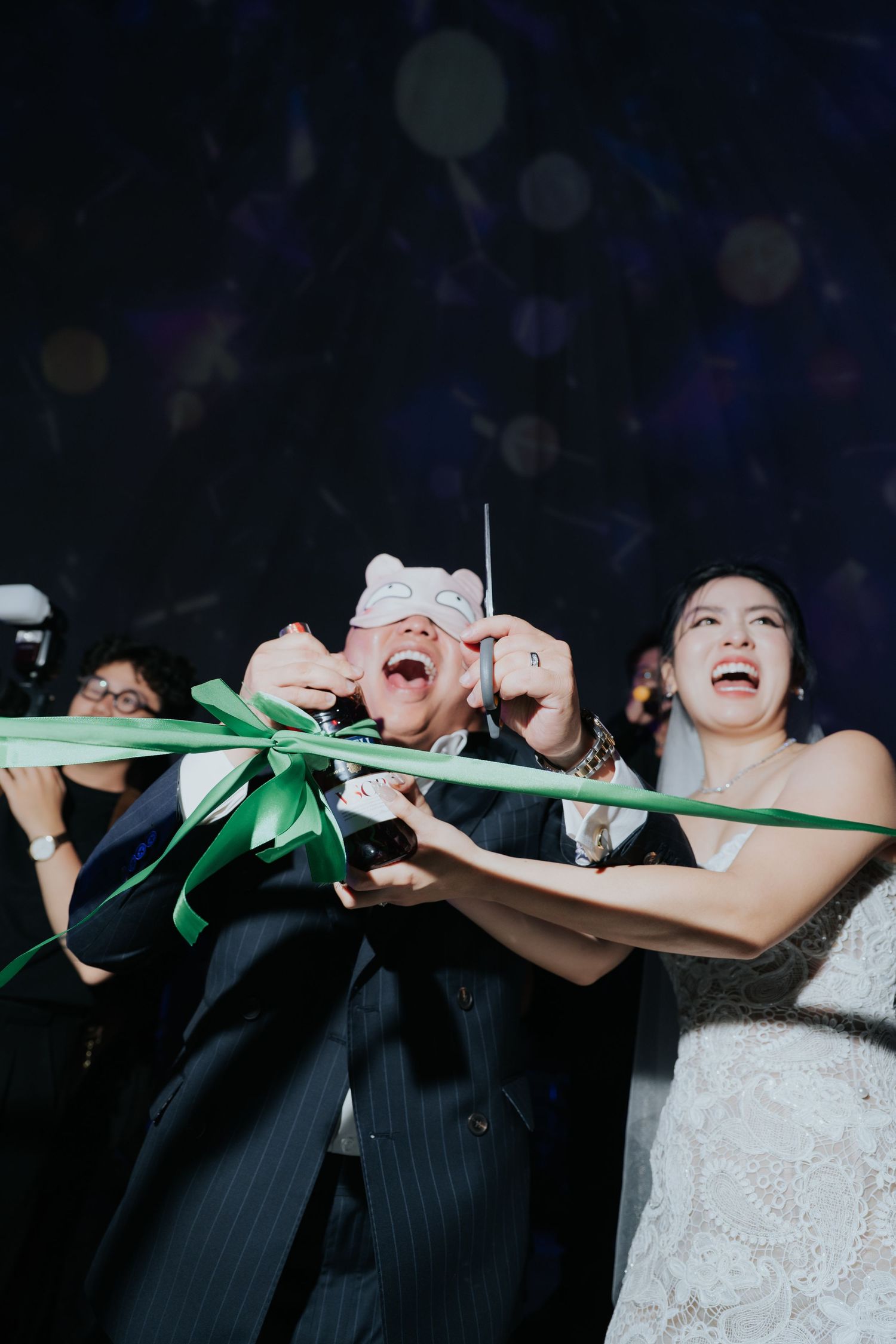 Bride and groom cutting green ribbon at wedding reception, wearing fun masks, laughing joyfully with guests celebrating behind them.