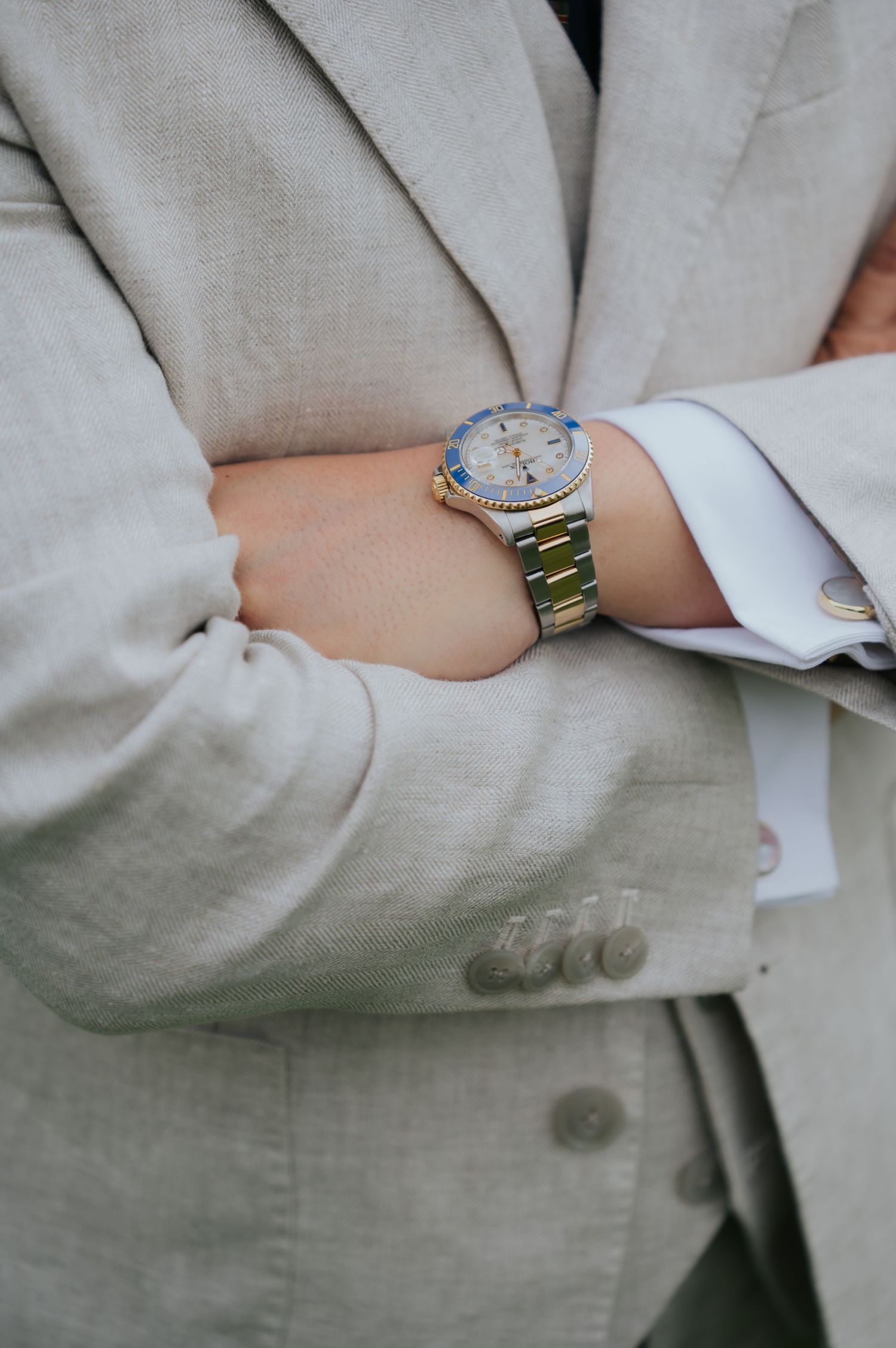 Man in beige linen suit wearing a gold Rolex watch with blue bezel, arms crossed, white dress shirt with cufflinks.