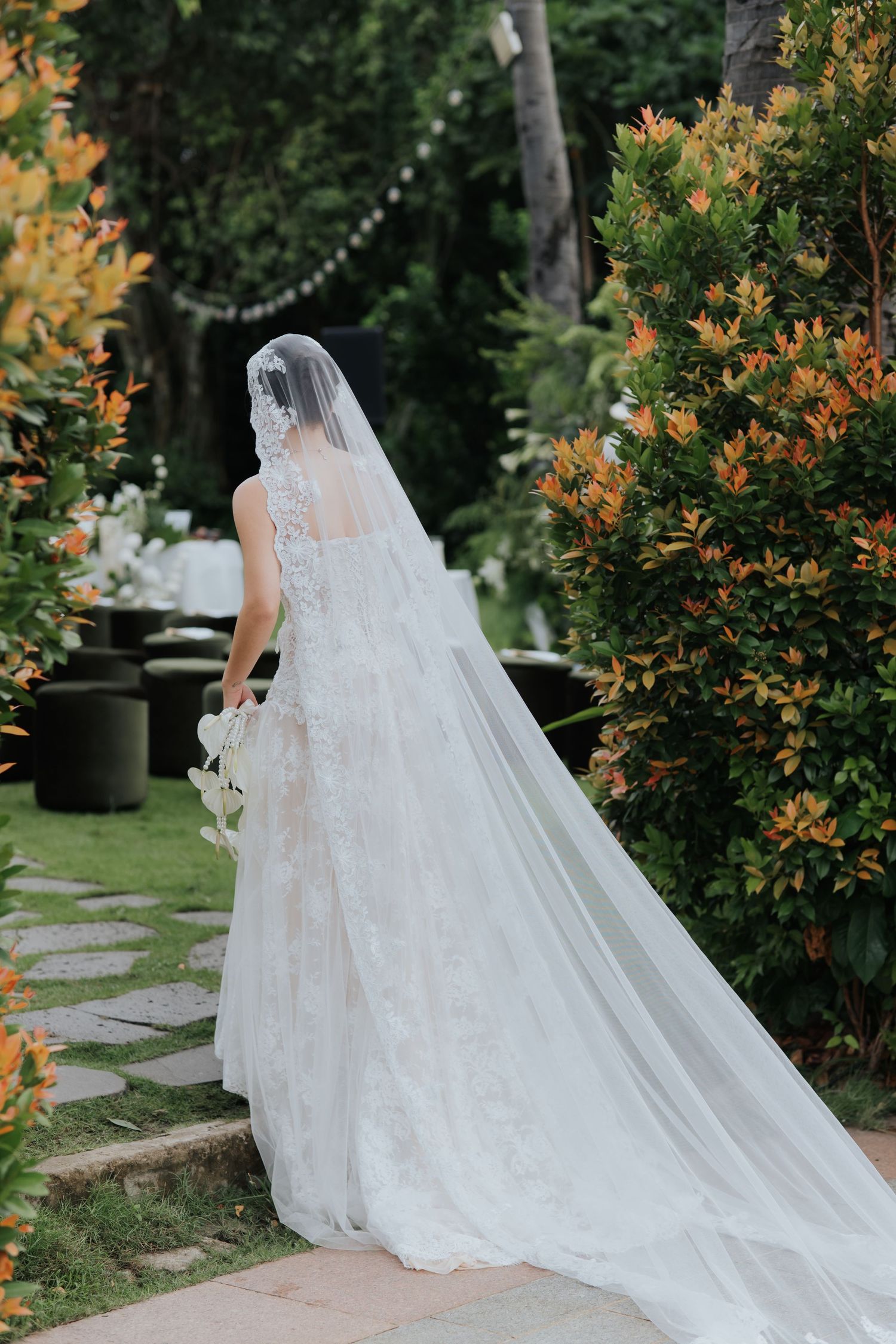 Bride in lace wedding gown with long veil walks through lush garden toward outdoor reception venue.