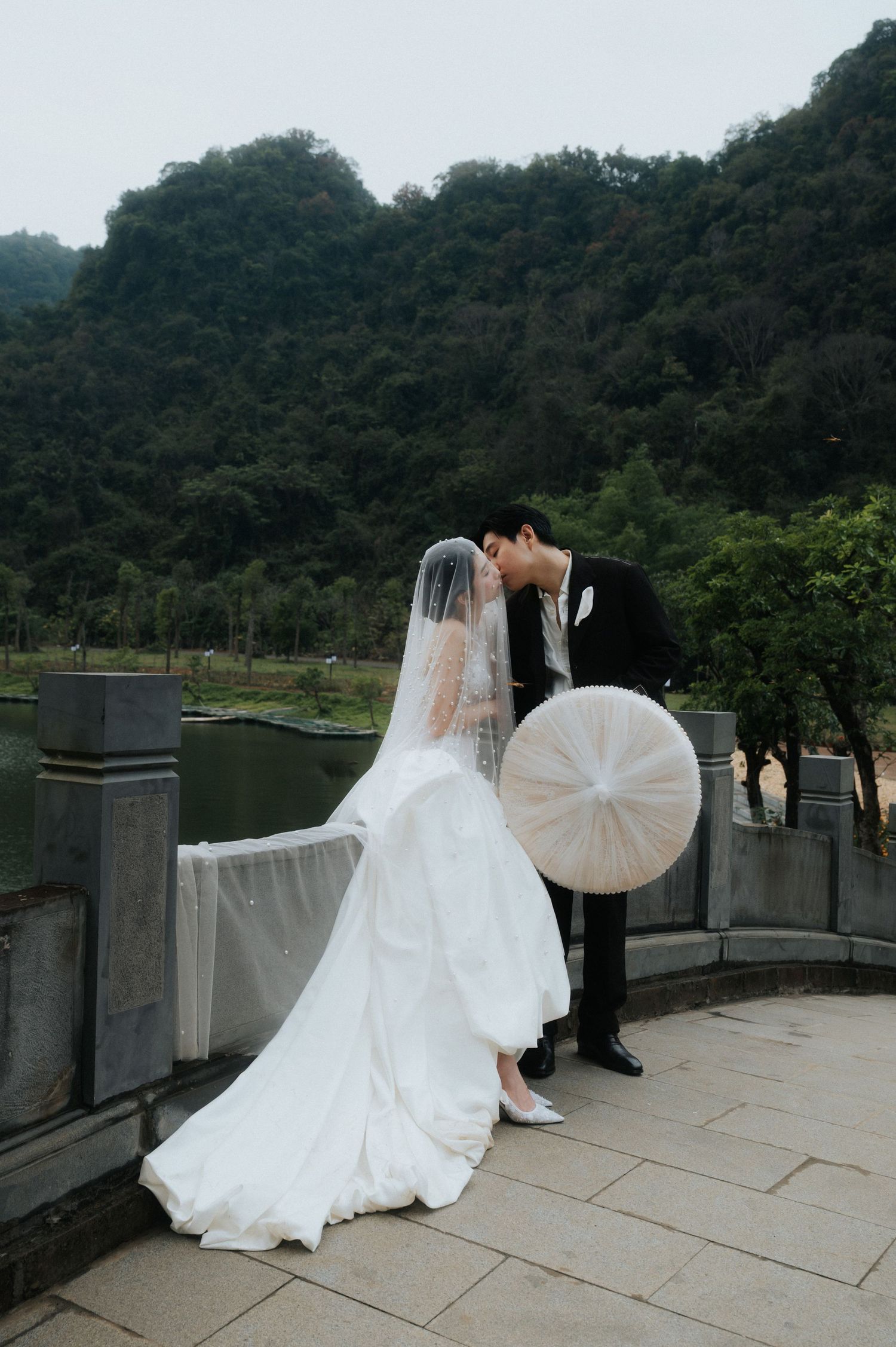 A bride and groom share a romantic kiss beside a serene lake, surrounded by lush green mountains, as she holds a white parasol.