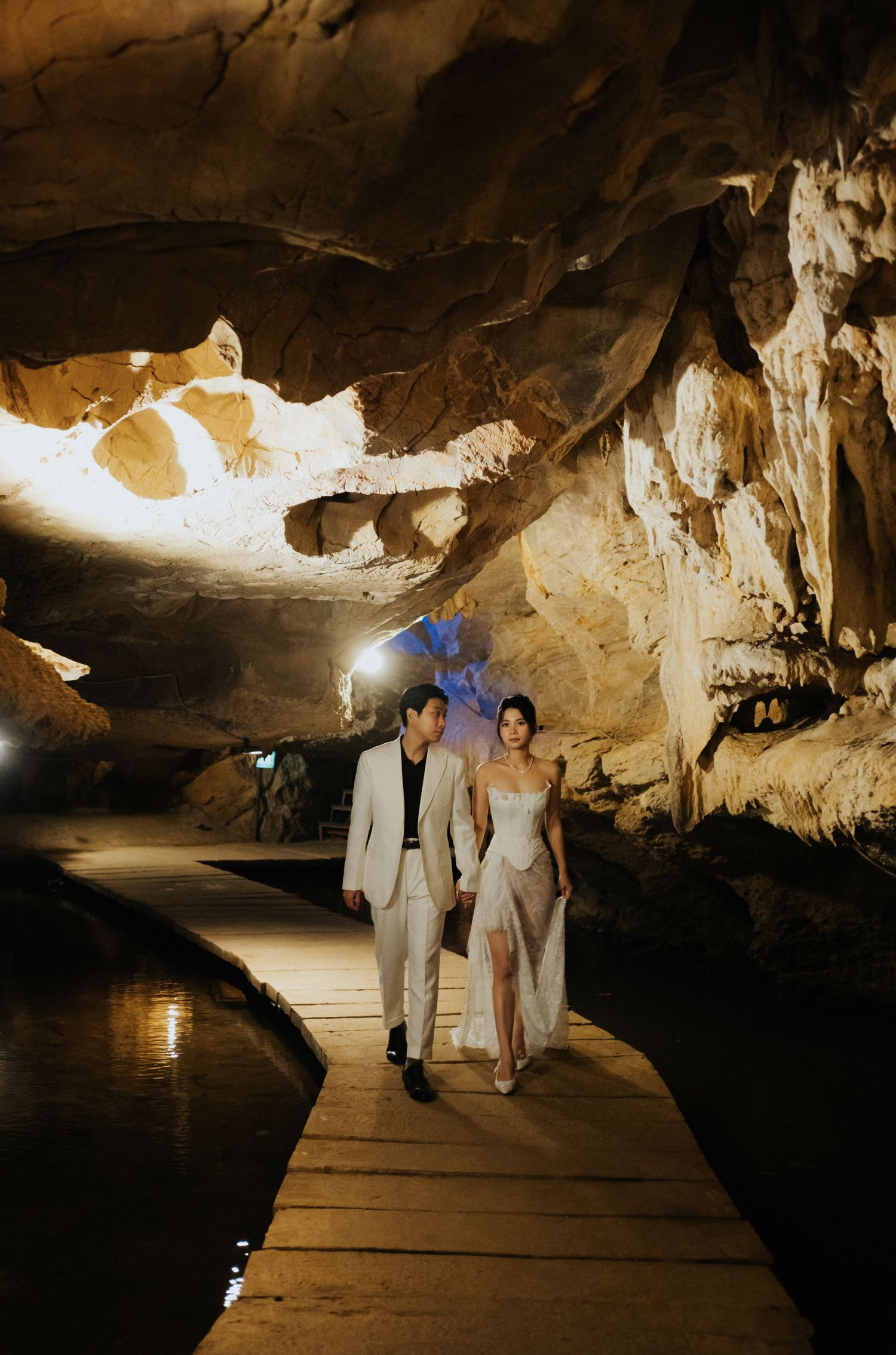 Couple in elegant attire walking through a stunning illuminated cave on a wooden boardwalk for a pre-wedding photoshoot.