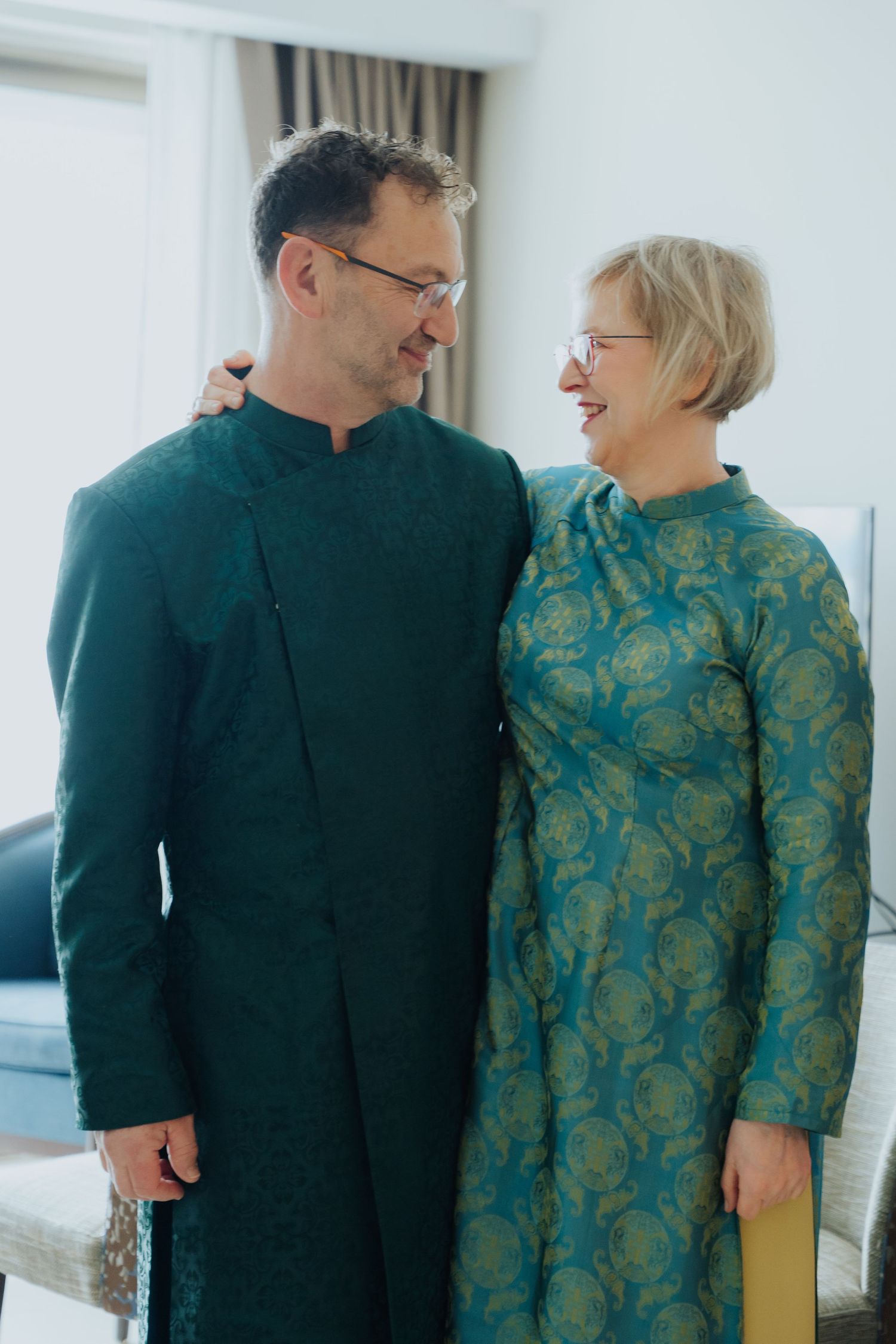 Couple in matching teal green traditional outfits smiling at each other in an elegant indoor setting.