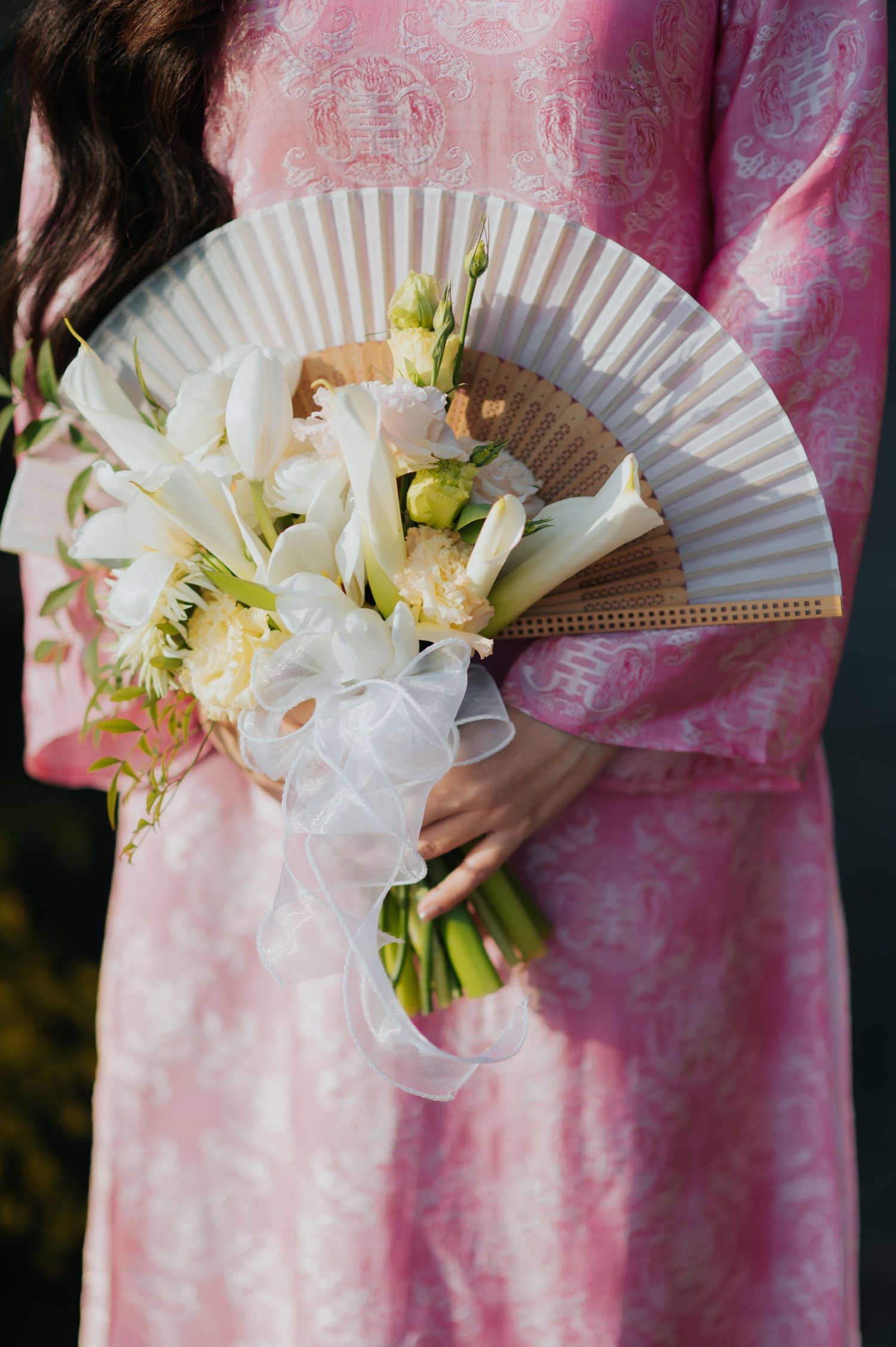 Woman in pink floral dress holding white lily bouquet with decorative hand fan and sheer ribbon.