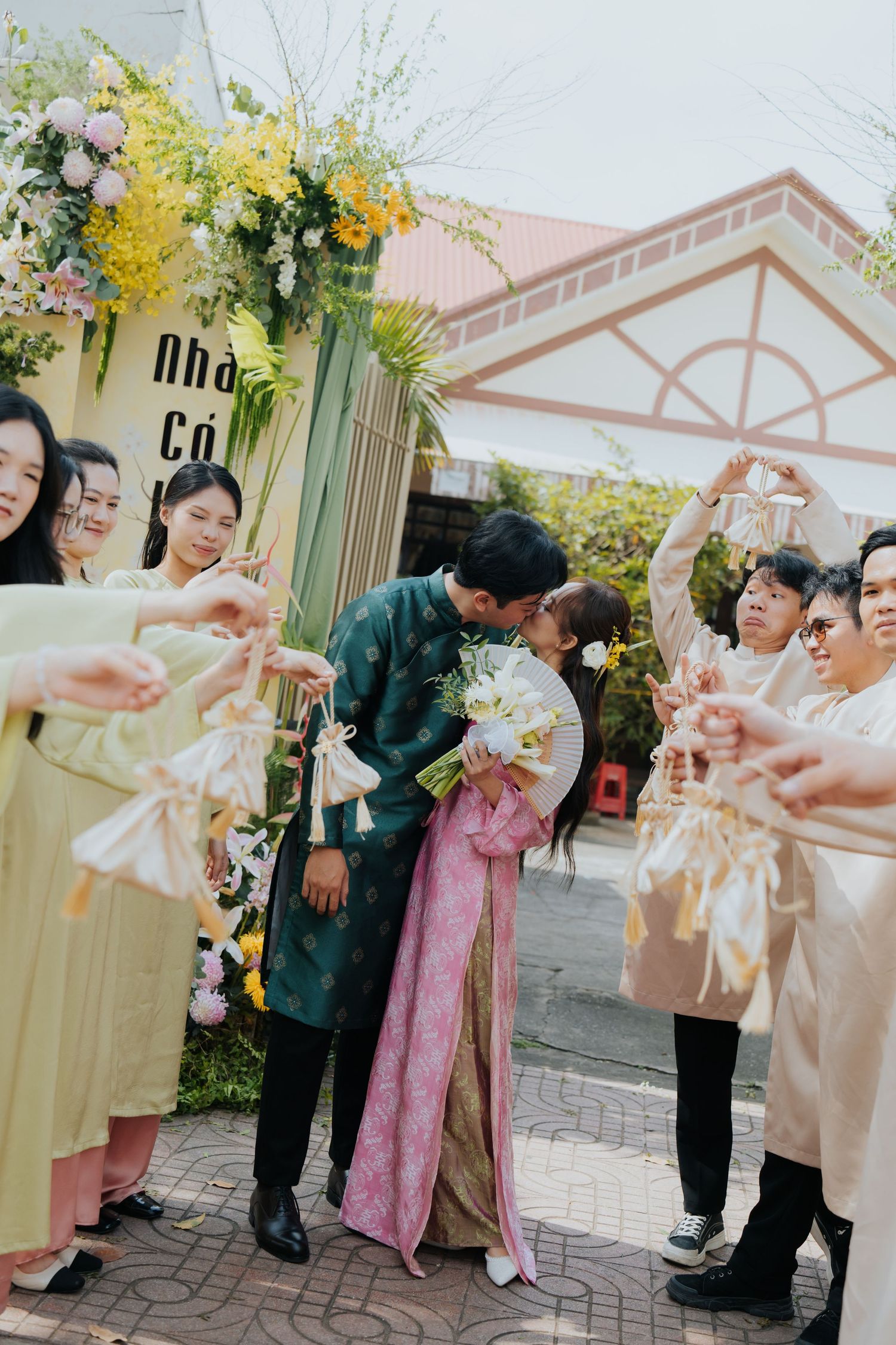 Vietnamese couple kissing at traditional wedding ceremony, surrounded by guests in ao dai attire holding floral decorations.