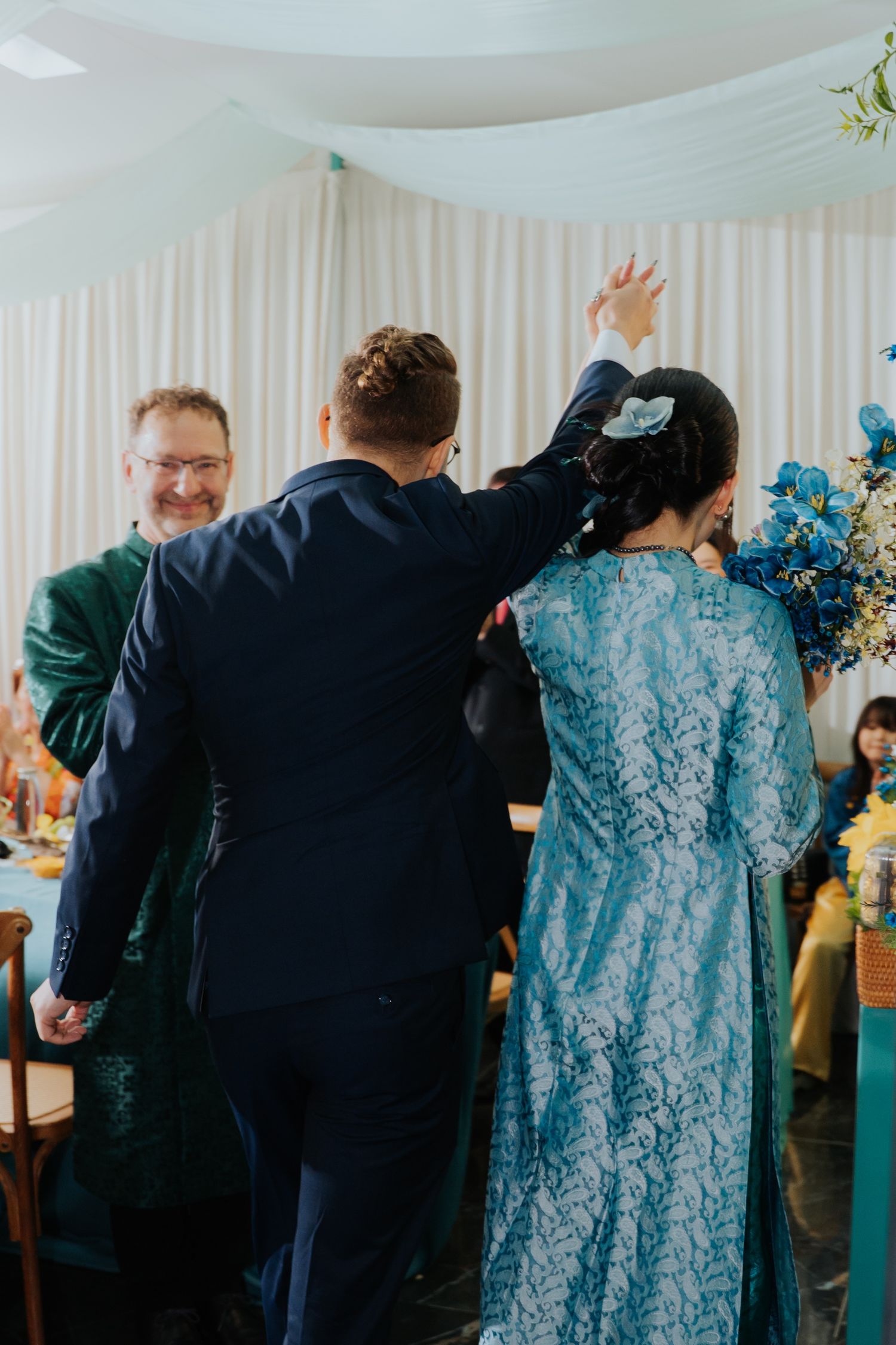 Groom in navy suit walks with woman in teal lace dress at elegant wedding reception with floral decor.