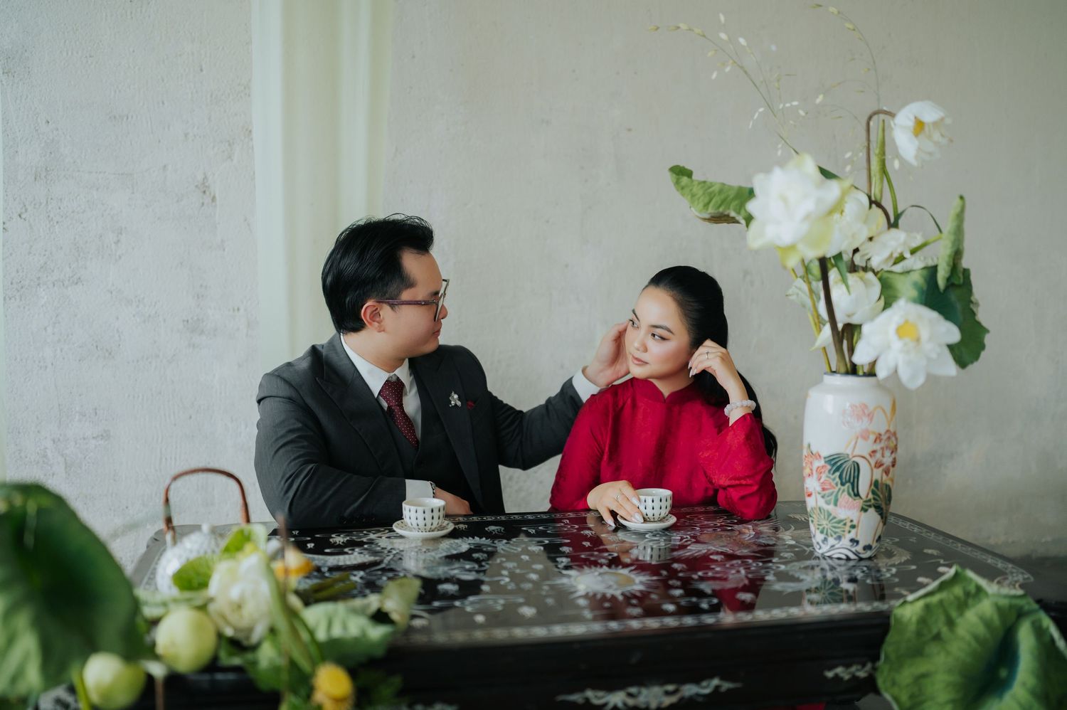 A couple in traditional Vietnamese attire sit at a table with tea cups and flowers, sharing an intimate moment.