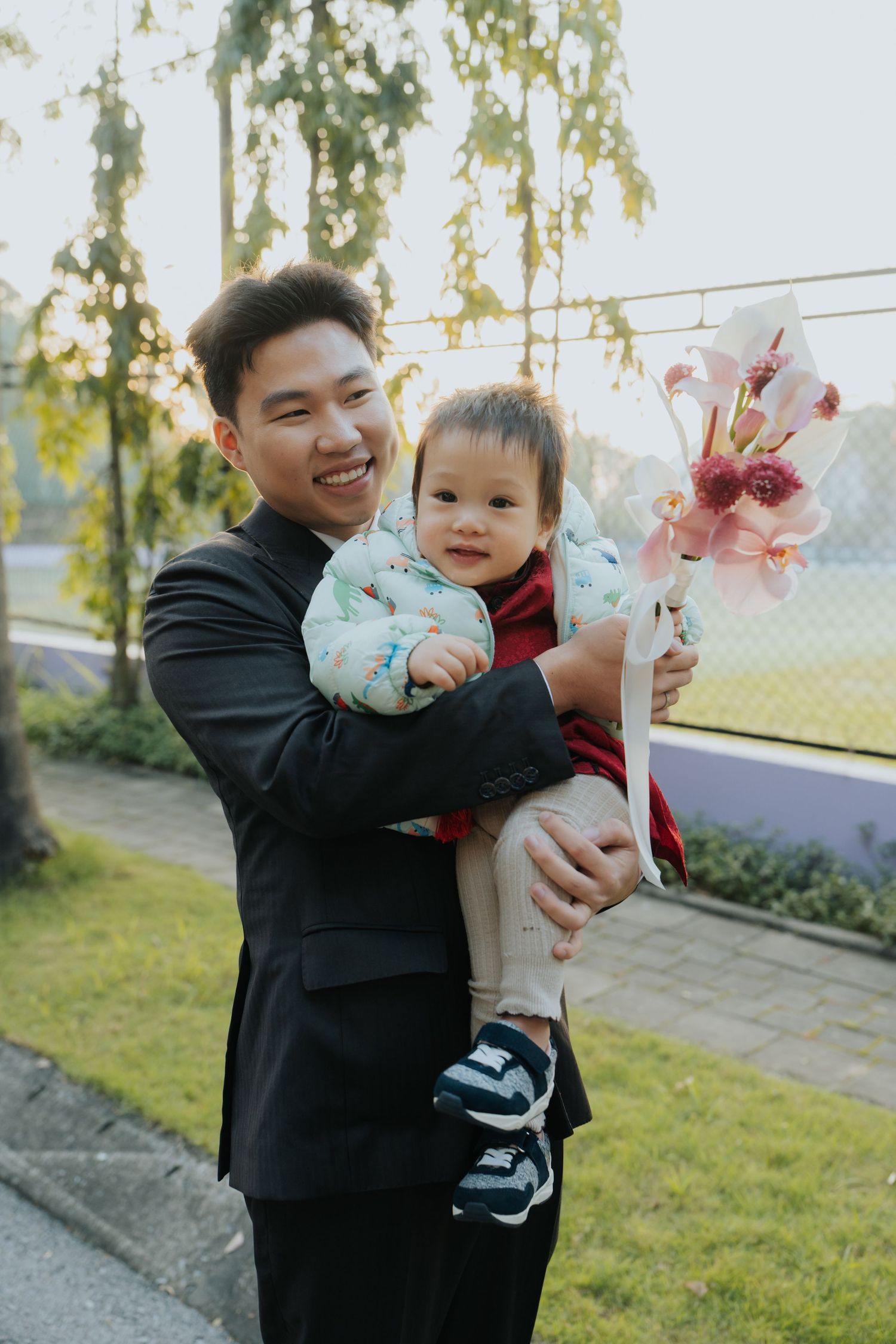 Smiling Asian man in black suit holding a baby and pink flower bouquet outdoors at sunset.