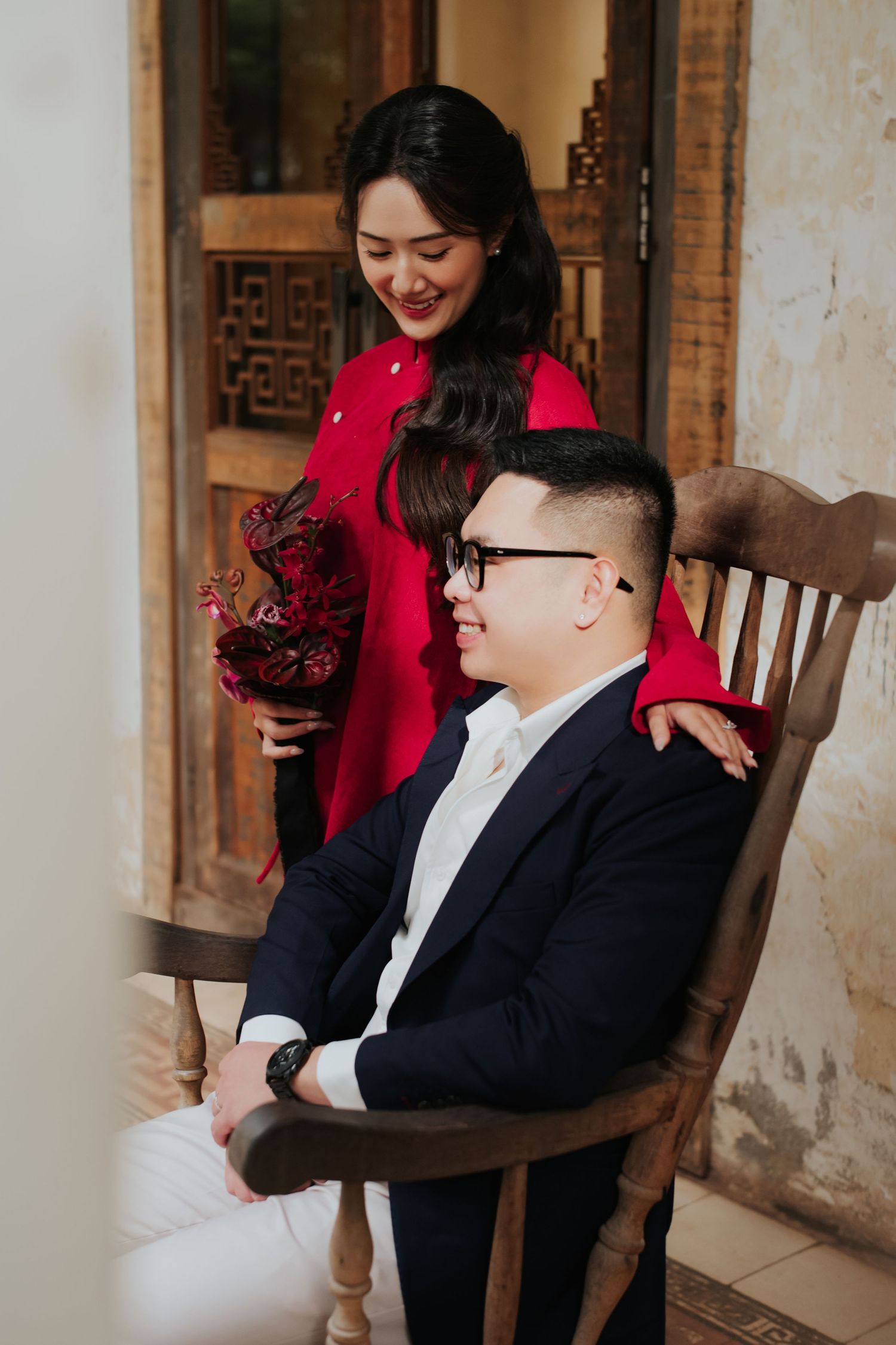 Couple in elegant attire, woman in red ao dai holding flowers stands behind man in navy suit seated in wooden chair.