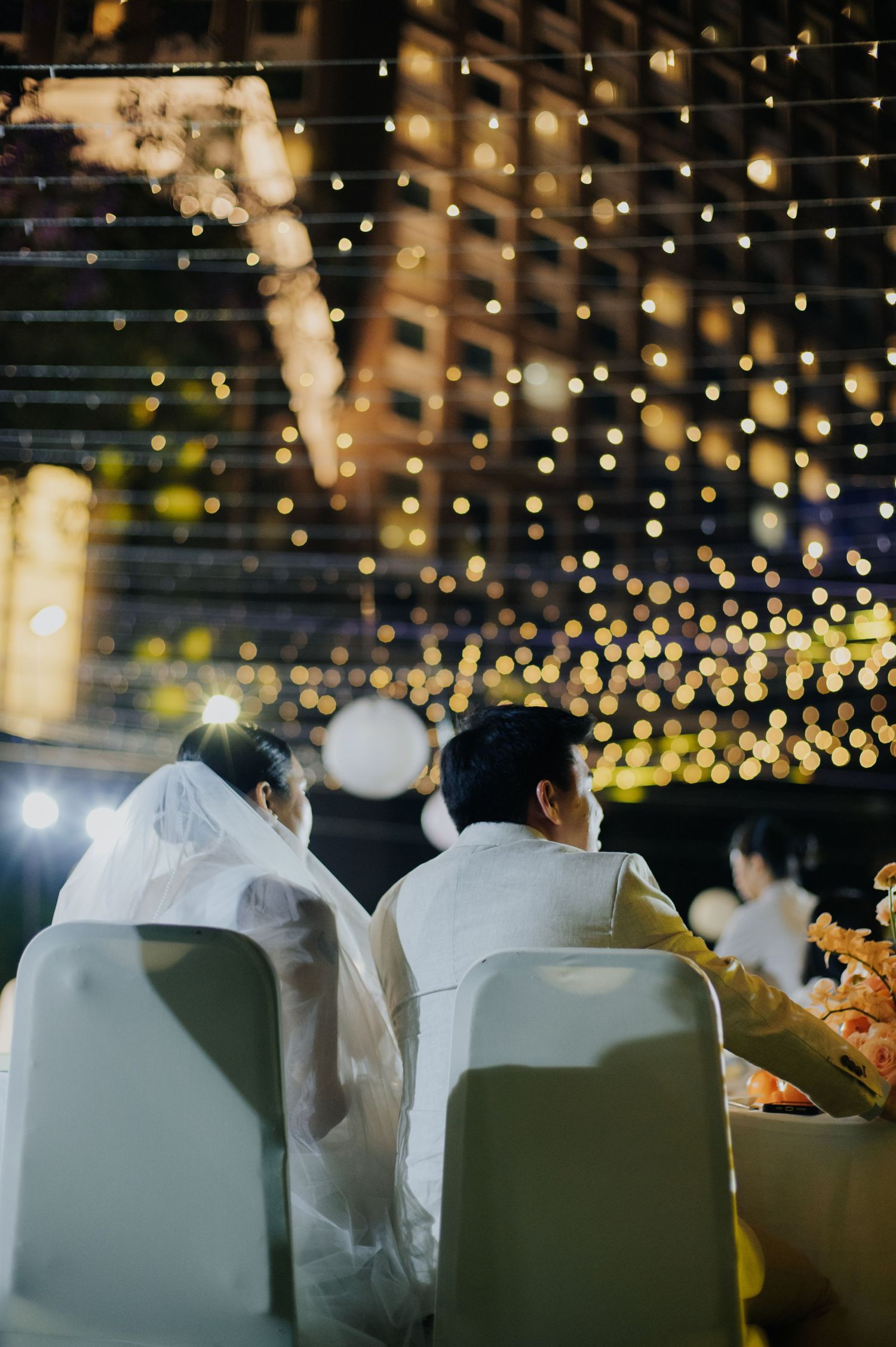 Couple in wedding attire seated at an outdoor evening reception with bokeh string lights glowing in the background.