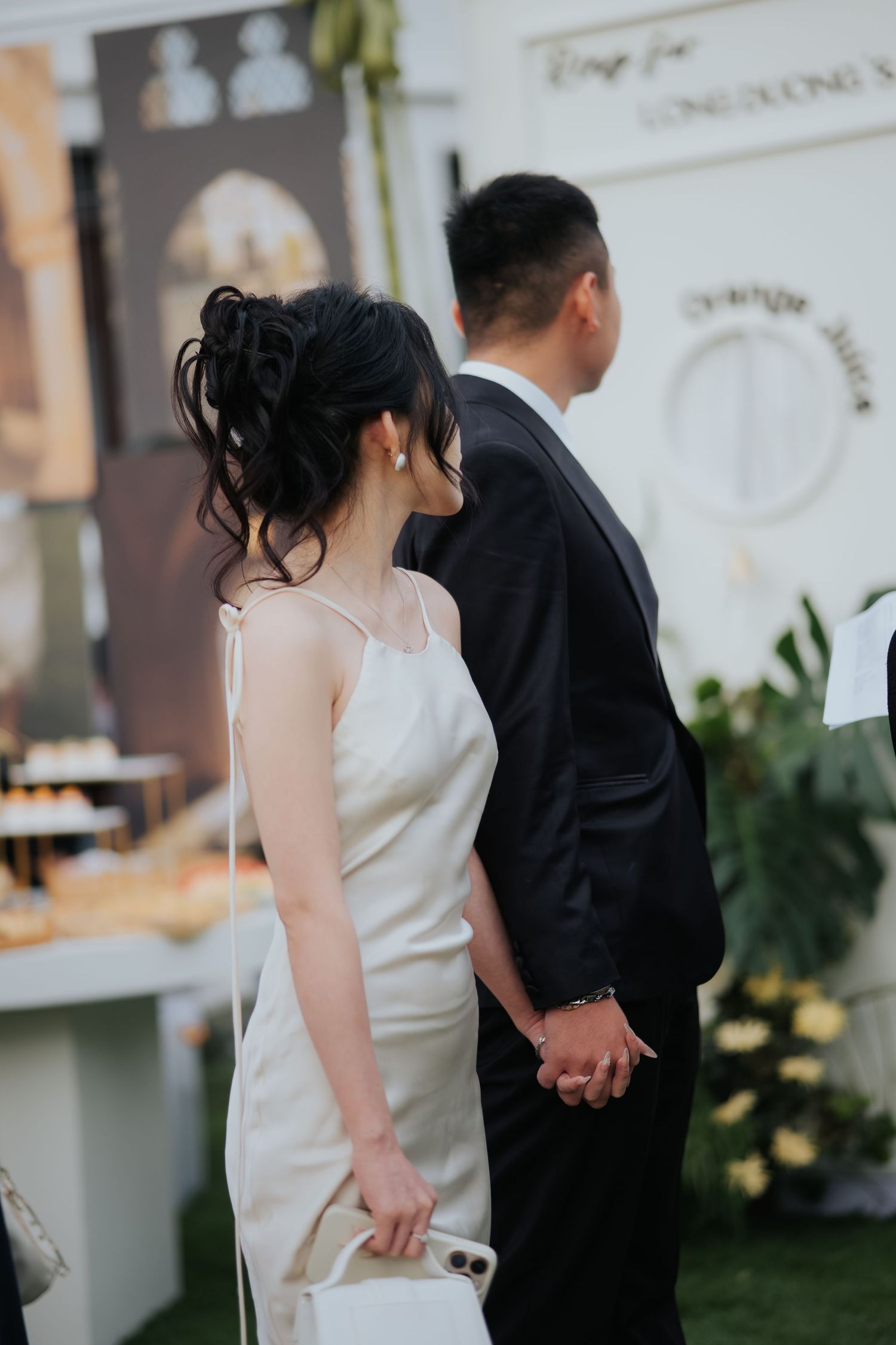A bride in a white satin slip dress holds hands with a groom in a black suit at an elegant wedding venue.