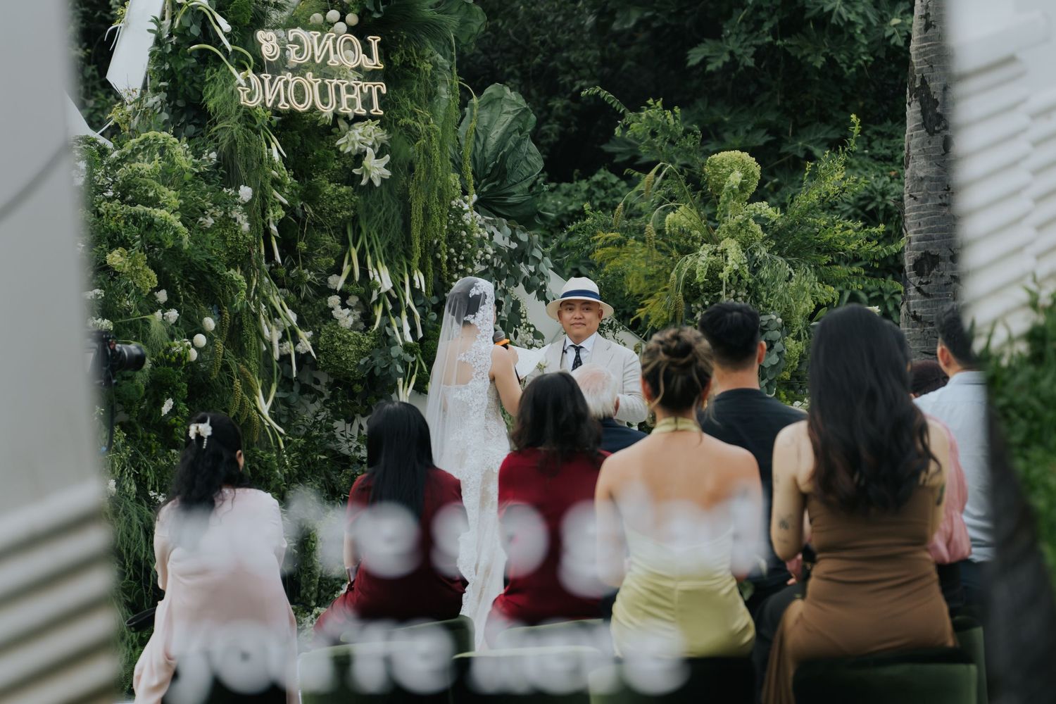 Outdoor wedding ceremony with bride and groom at floral arch, guests seated on chairs surrounded by lush tropical greenery.