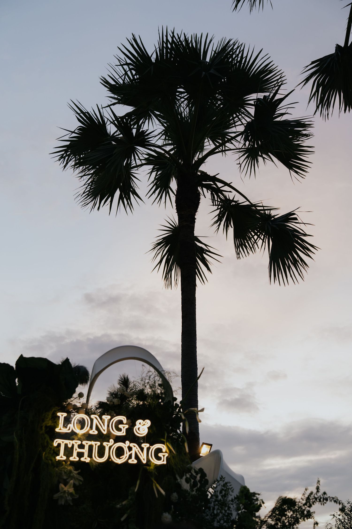 Illuminated 'Long & Thuong' wedding sign glowing at dusk beneath a tall silhouetted palm tree against a twilight sky.