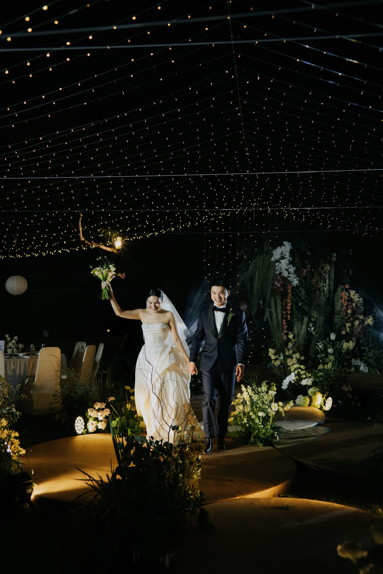 Bride and groom walk down aisle under twinkling fairy lights at elegant outdoor night wedding reception.