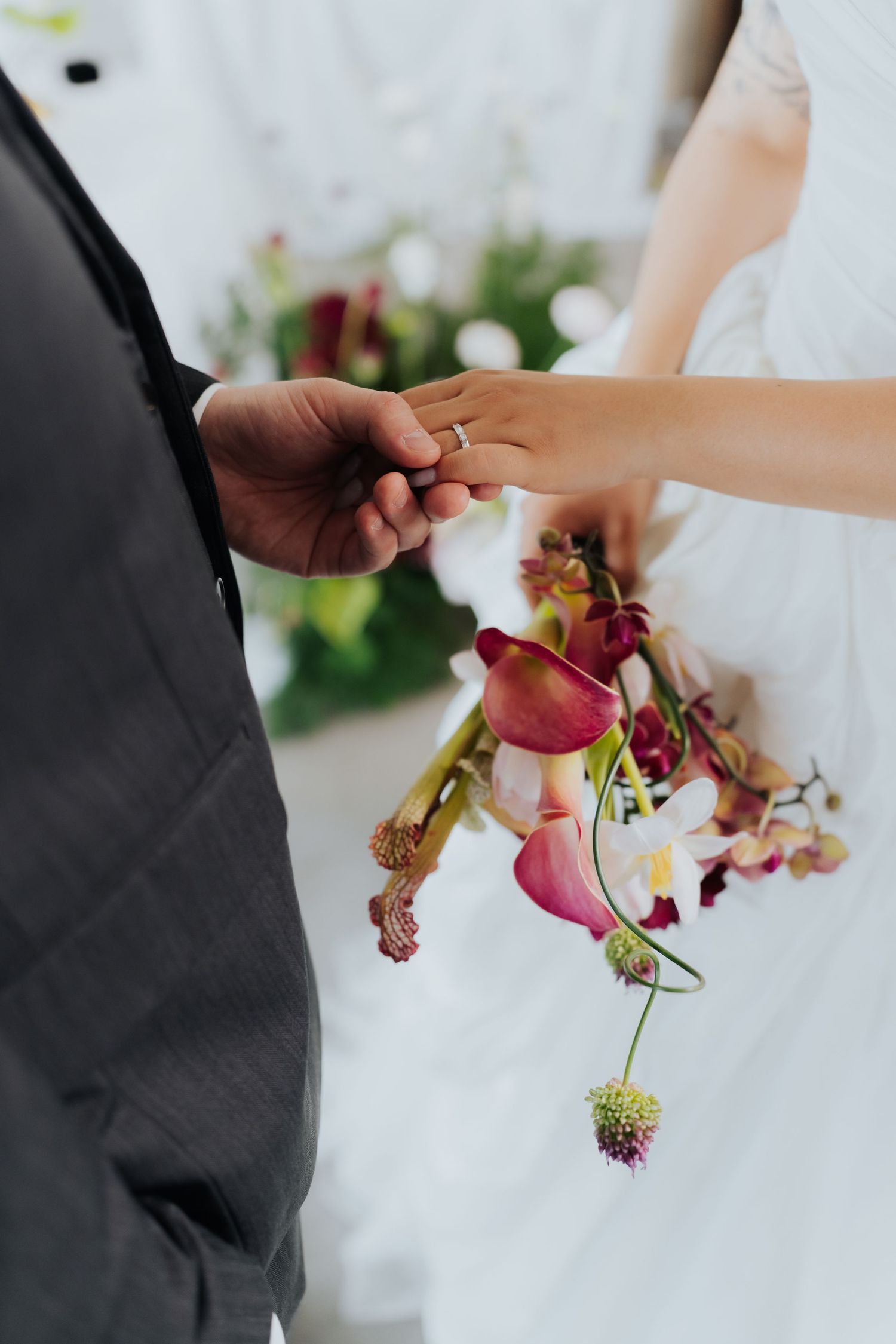 Bride placing a wedding ring on groom's finger during ceremony, holding a bouquet of pink calla lilies and orchids.