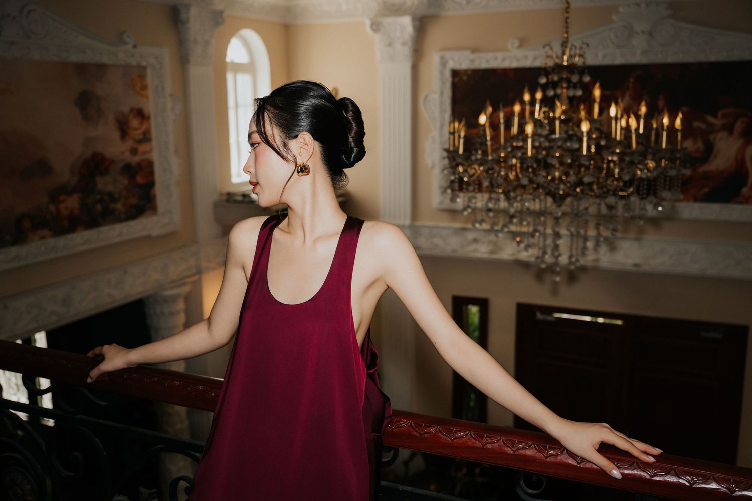 A young woman in a burgundy dress stands by a railing in an elegant room with a crystal chandelier.