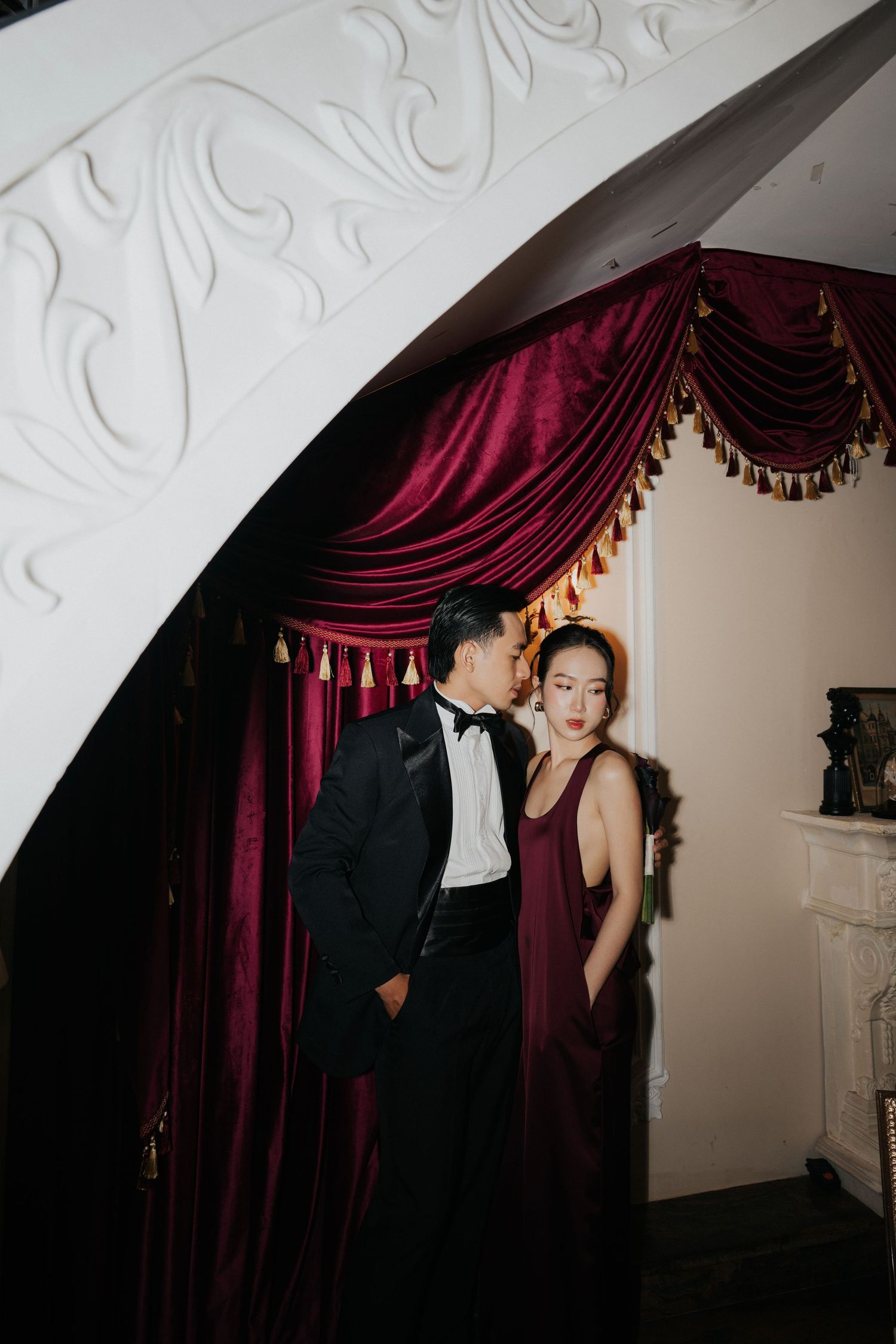 Elegant couple in formal attire posing by a grand staircase with rich burgundy velvet drapes.