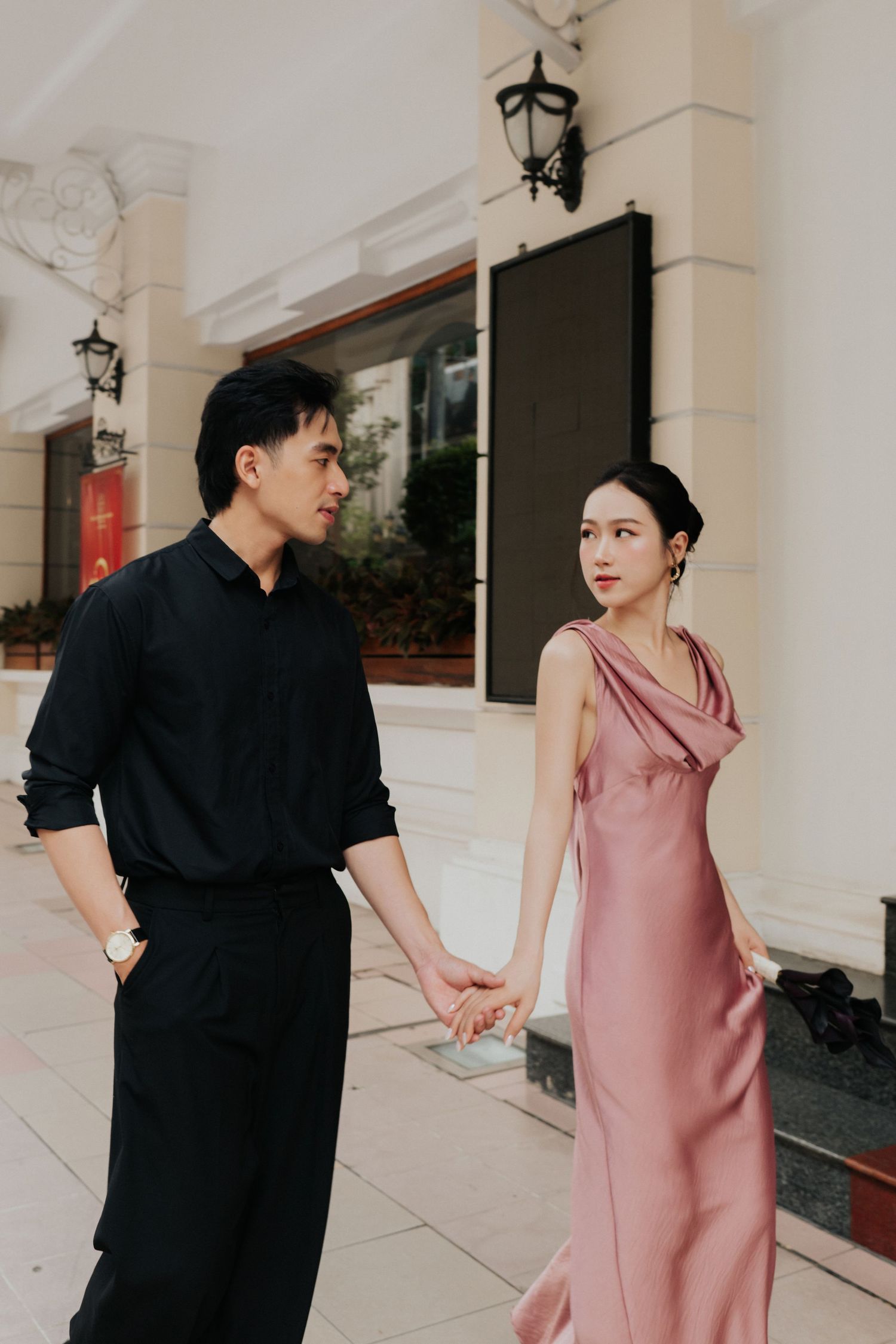 A couple holds hands outdoors, man in black outfit, woman in elegant pink satin dress, against a classic white building.