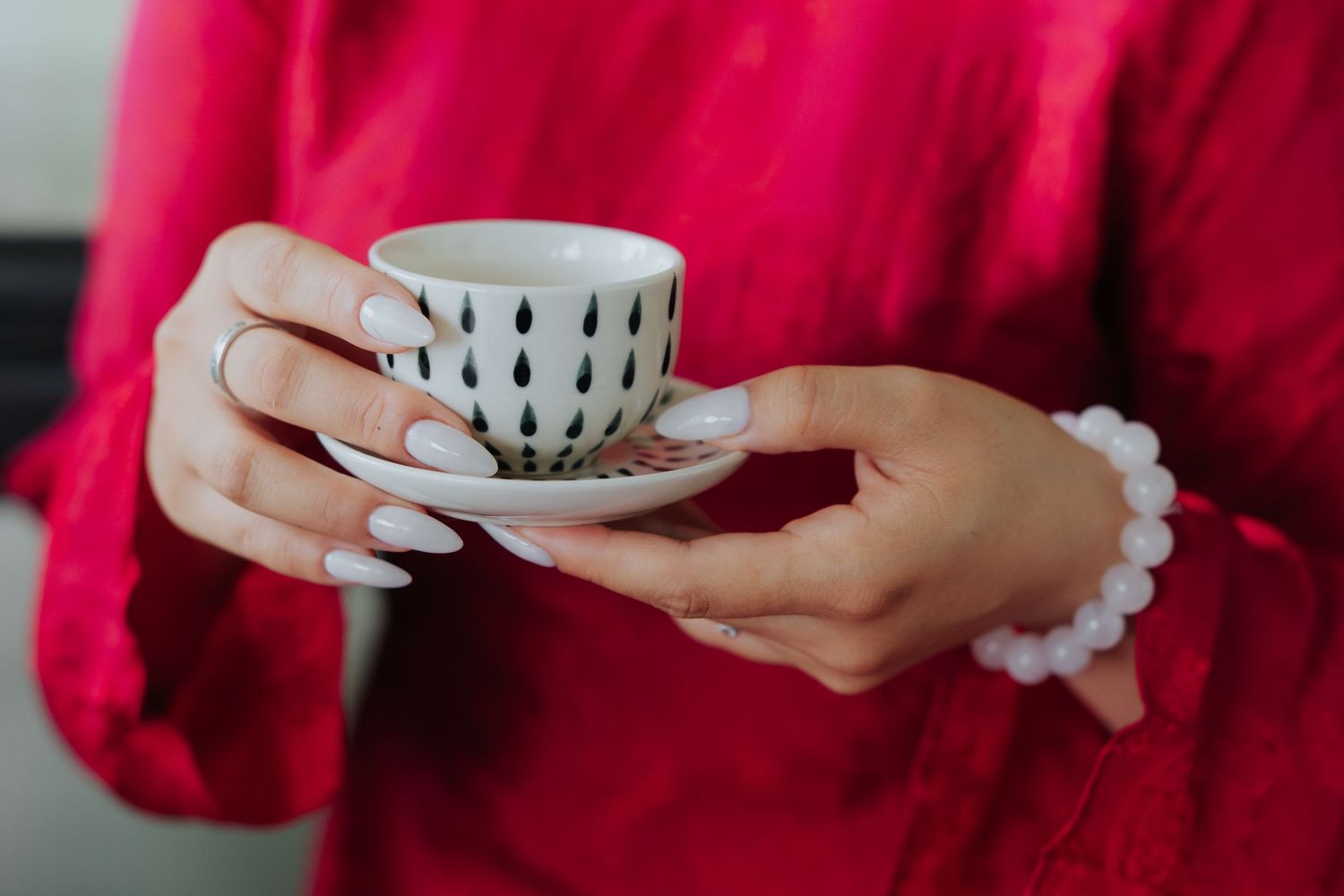Woman in red blouse holding a white ceramic espresso cup and saucer with black raindrop pattern and pearl bracelet.