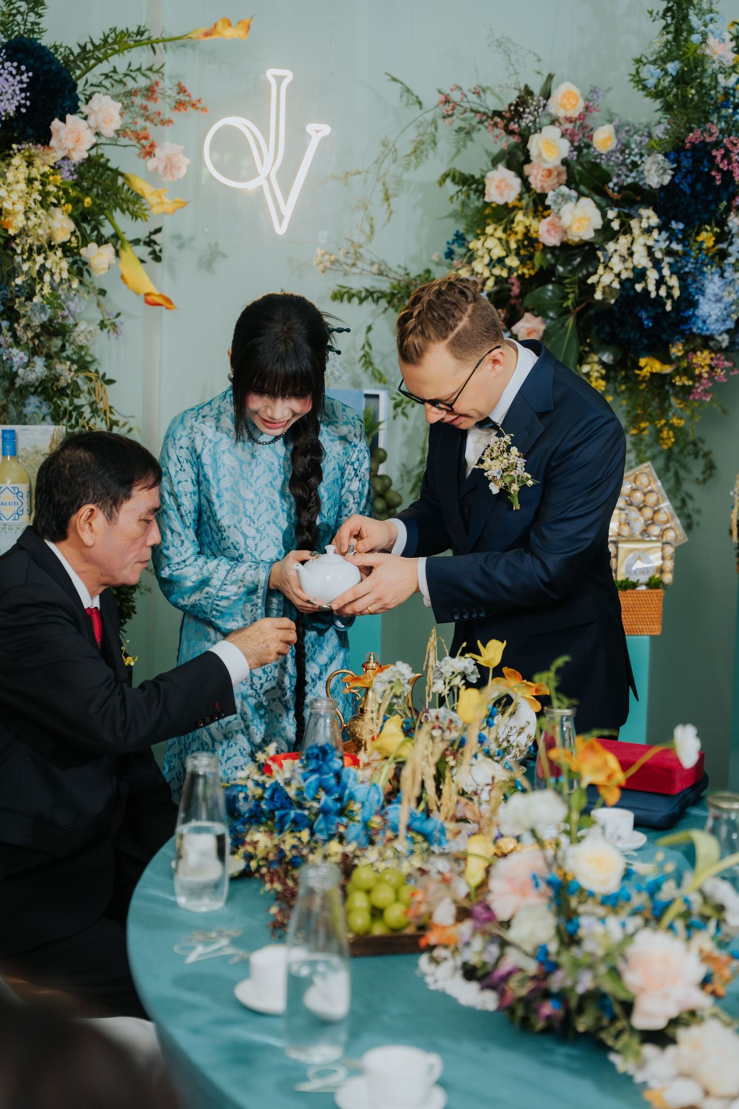 Couple in traditional Vietnamese wedding attire performing tea ceremony at floral-decorated table with monogram backdrop.