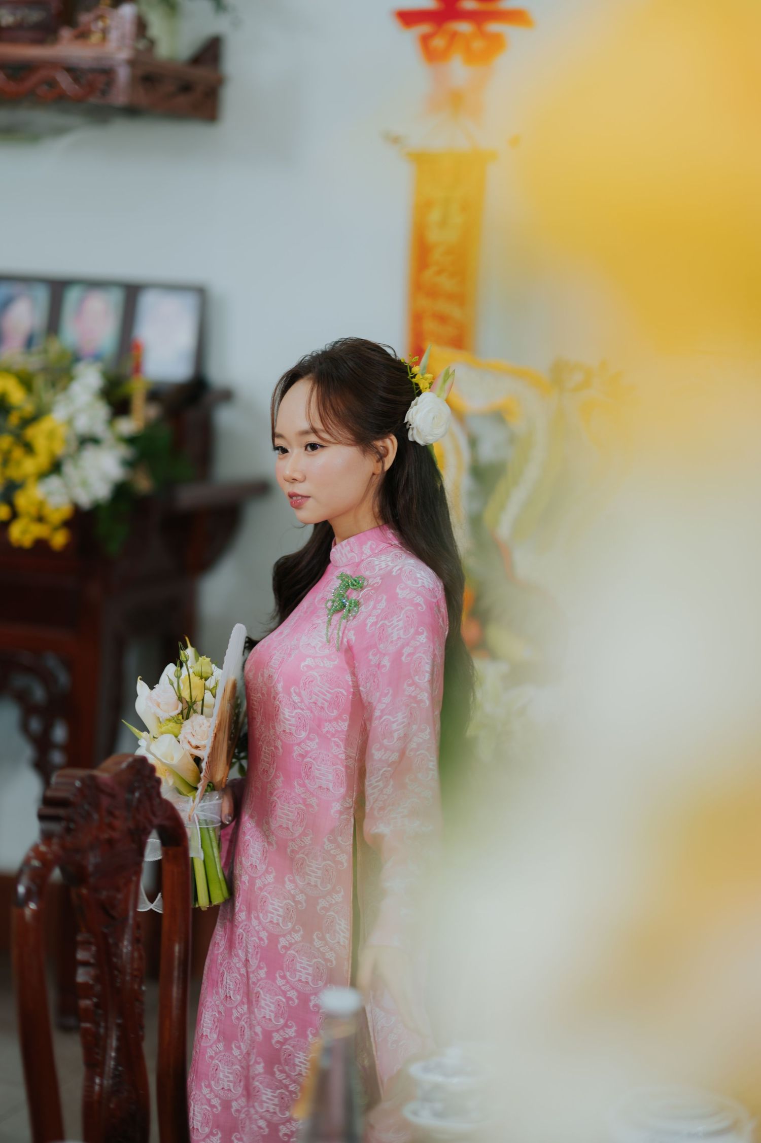 Young Vietnamese woman in pink floral ao dai holding white bouquet with hair flower at traditional ceremony.