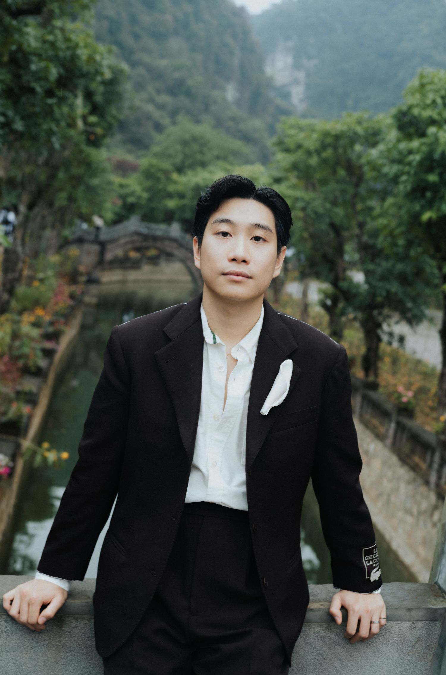 Young Asian man in black suit with white pocket square poses outdoors near a scenic river and lush green karst mountains.